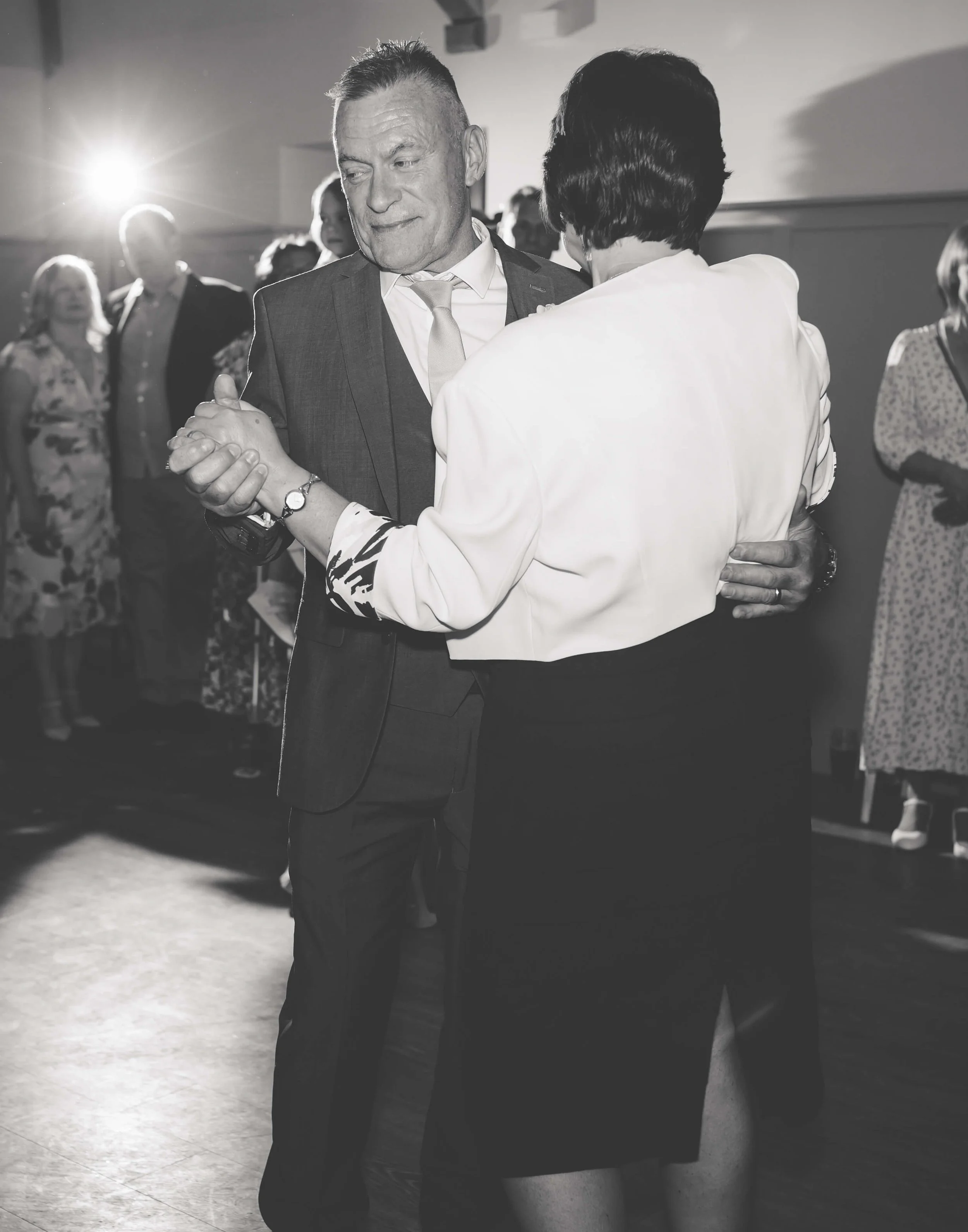 A black-and-white photo of a man and woman dancing together at a social event. The man is in a suit, and the woman is in a white blouse and black skirt. Other people are visible in the background.