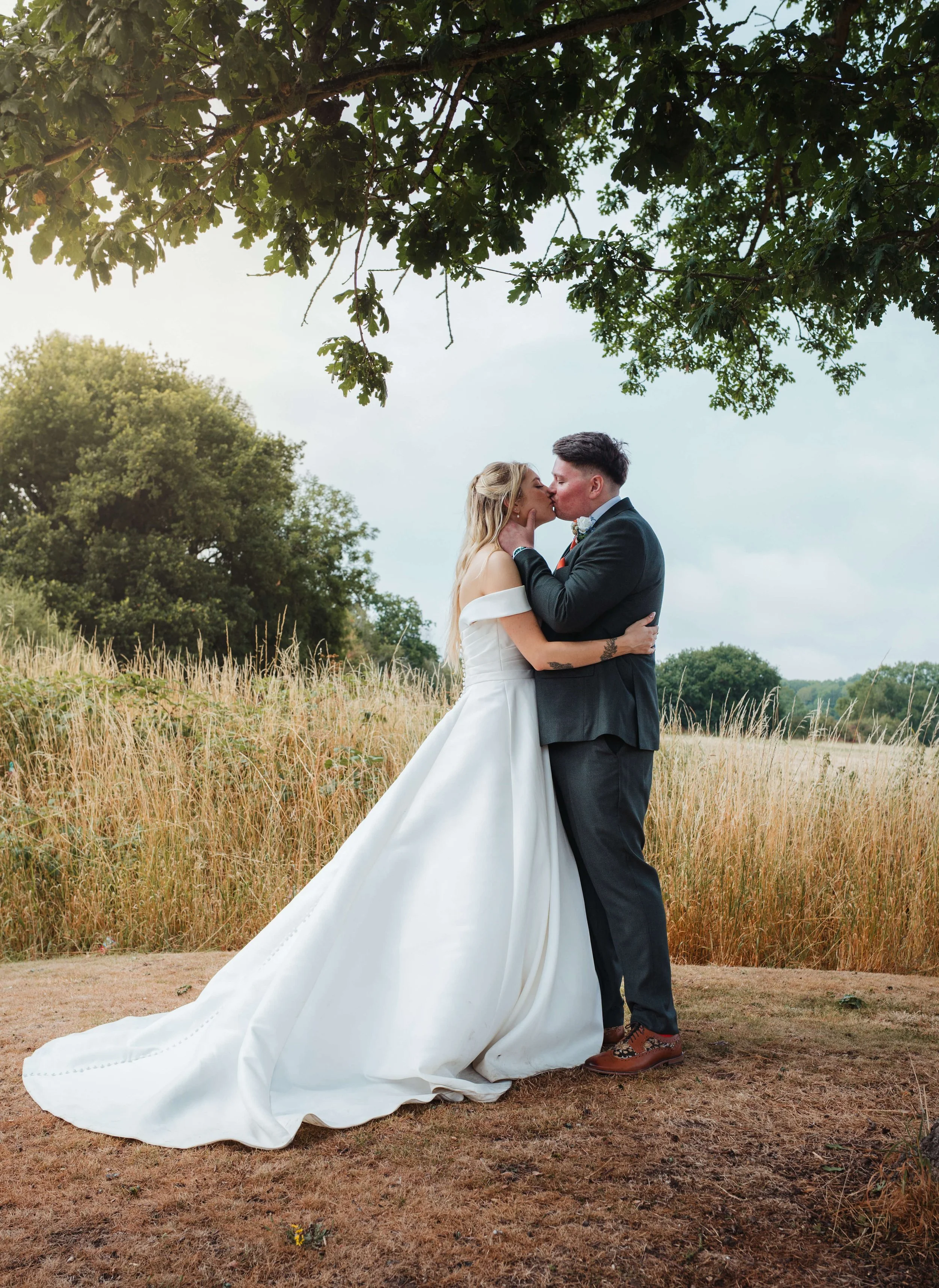 A bride and groom sharing a kiss outdoors during their wedding, standing on dirt with tall grass and trees in the background.