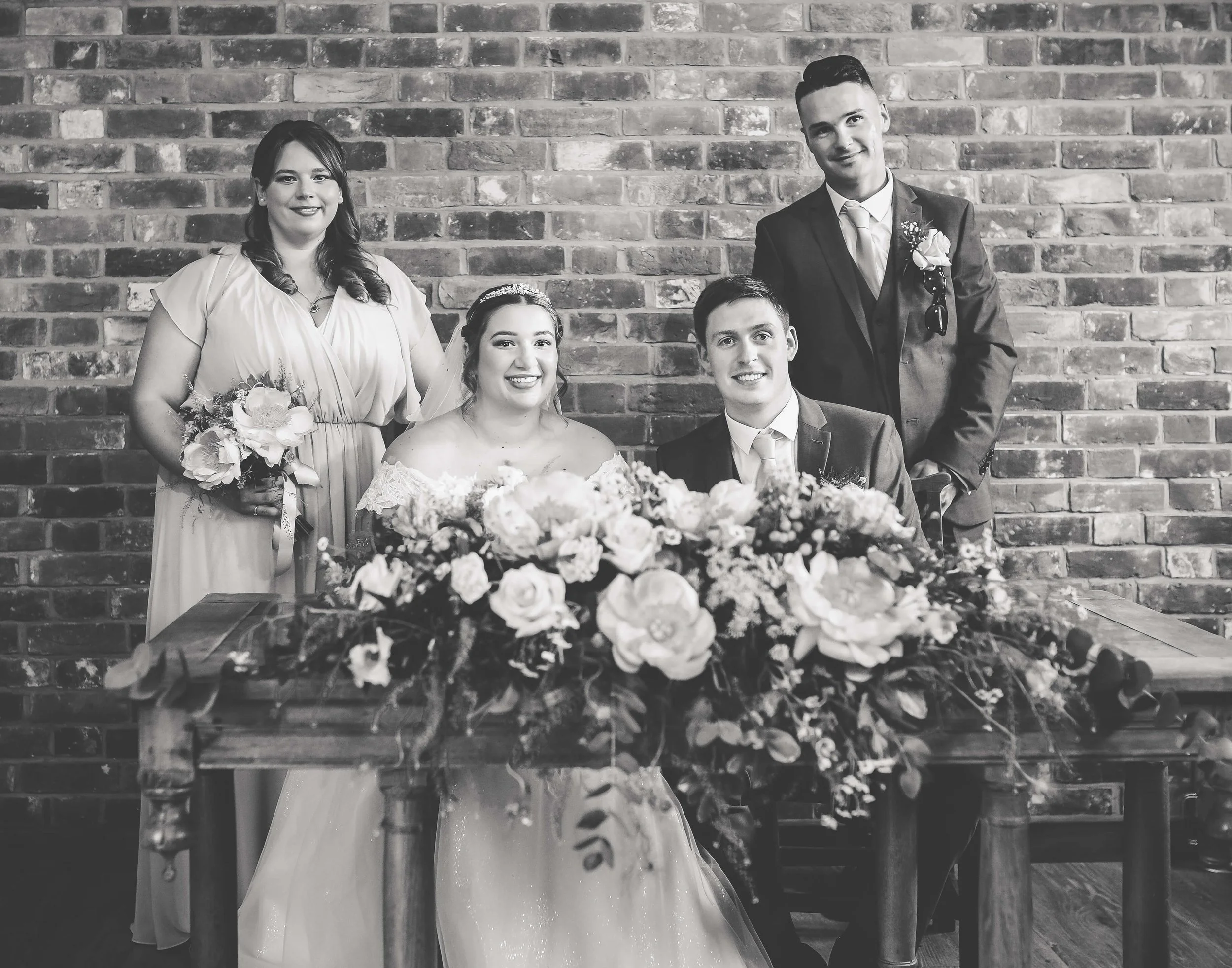 Black and white photo of a wedding party. The bride and groom are sitting at a wooden table with a large floral arrangement. Two women and one man stand behind them against a brick wall. The woman on the left holds a bouquet, the bride wears a veil a