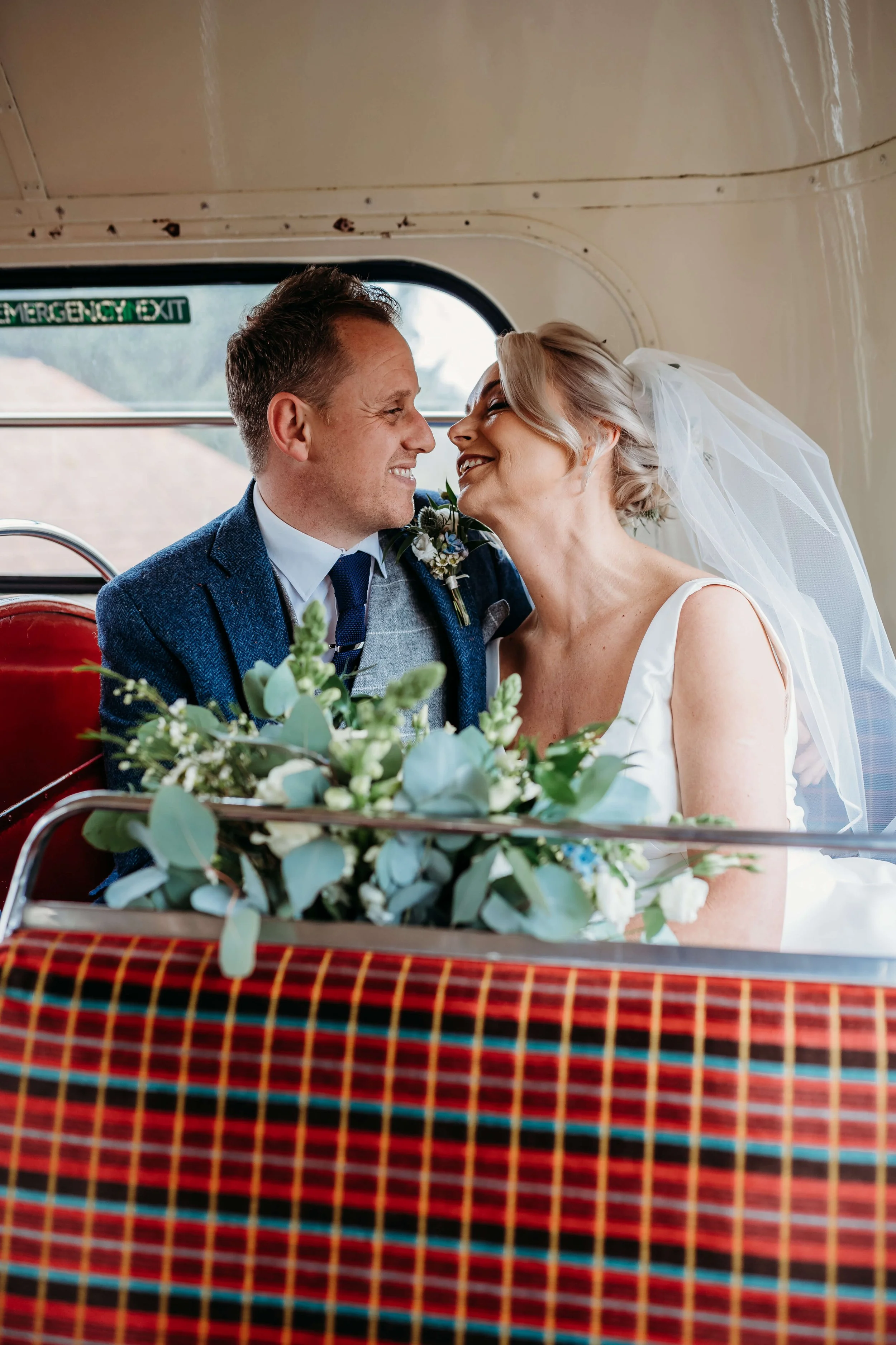 A bride and groom smiling and leaning close together on a bus, with a bouquet of flowers in front of them, celebrating their wedding.