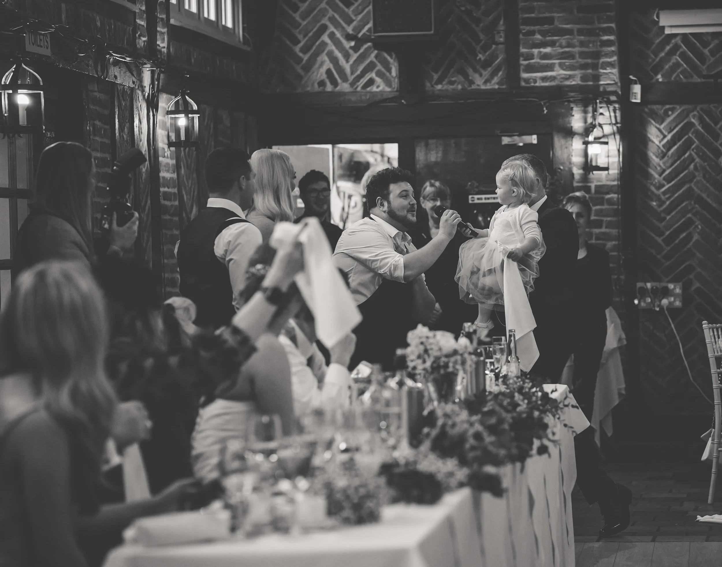 A black and white photo showing a wedding reception scene with a man holding a microphone for a little girl on a table, surrounded by people in formal attire inside a rustic venue with brick walls and chandeliers.