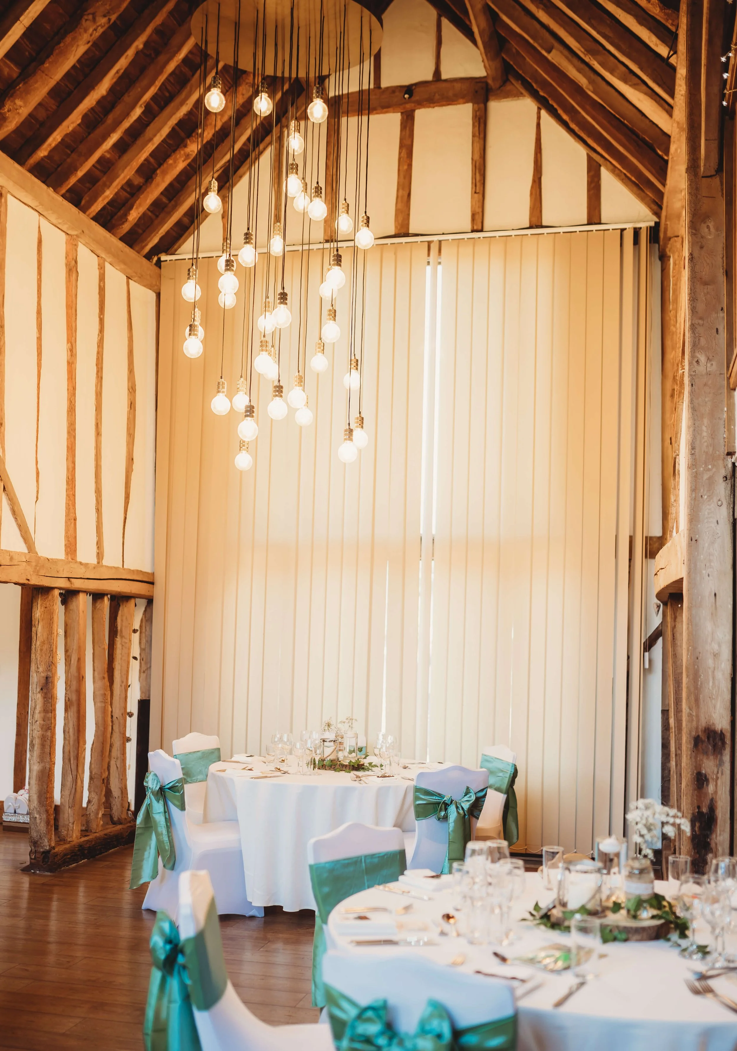 Elegant banquet hall with rustic wooden beams, a chandelier with multiple glowing bulbs, and round tables decorated with white tablecloths, floral centerpieces, and teal ribbon accents on chairs.