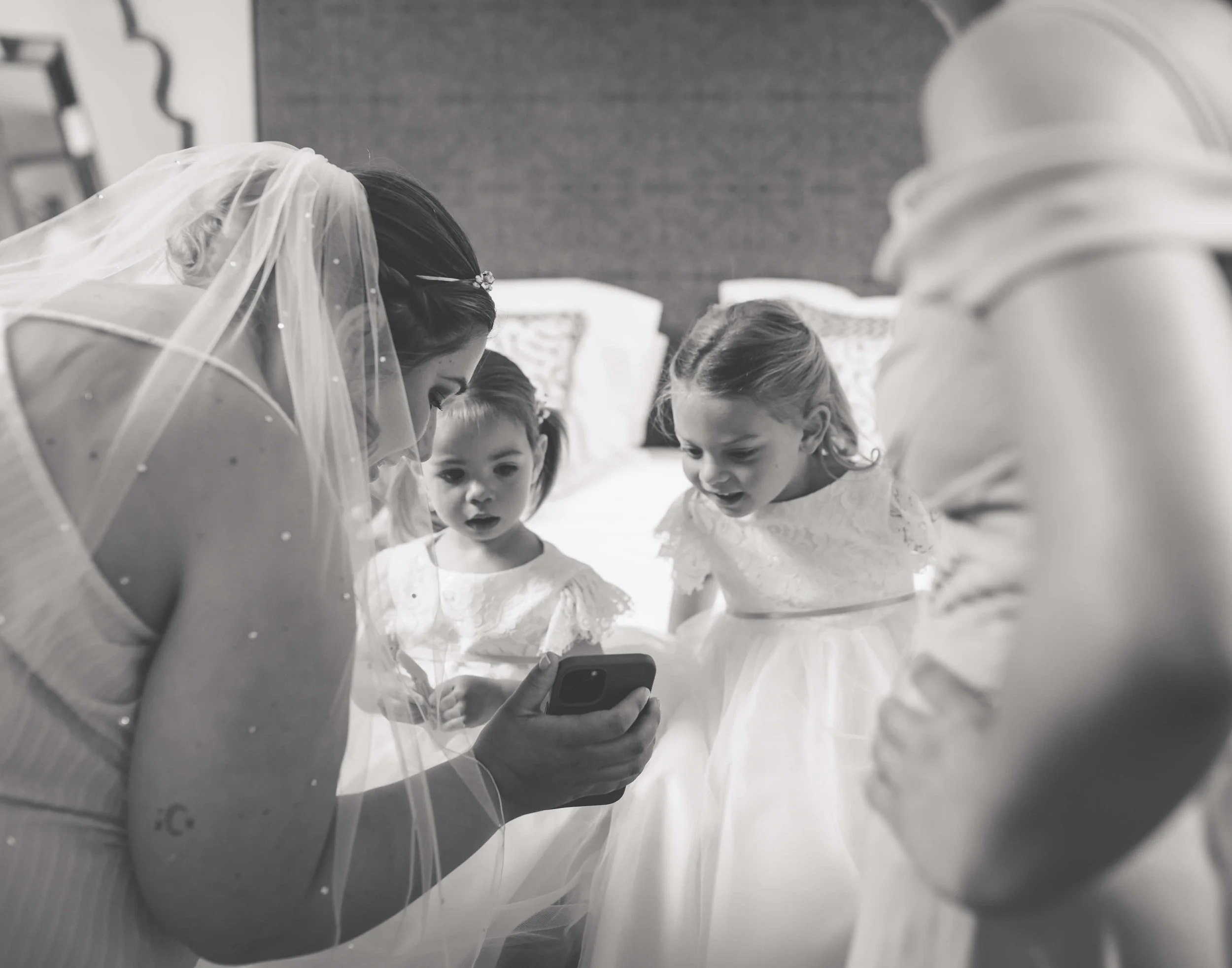 A bride with a veil and three young bridesmaids looking at a phone together in a bedroom.