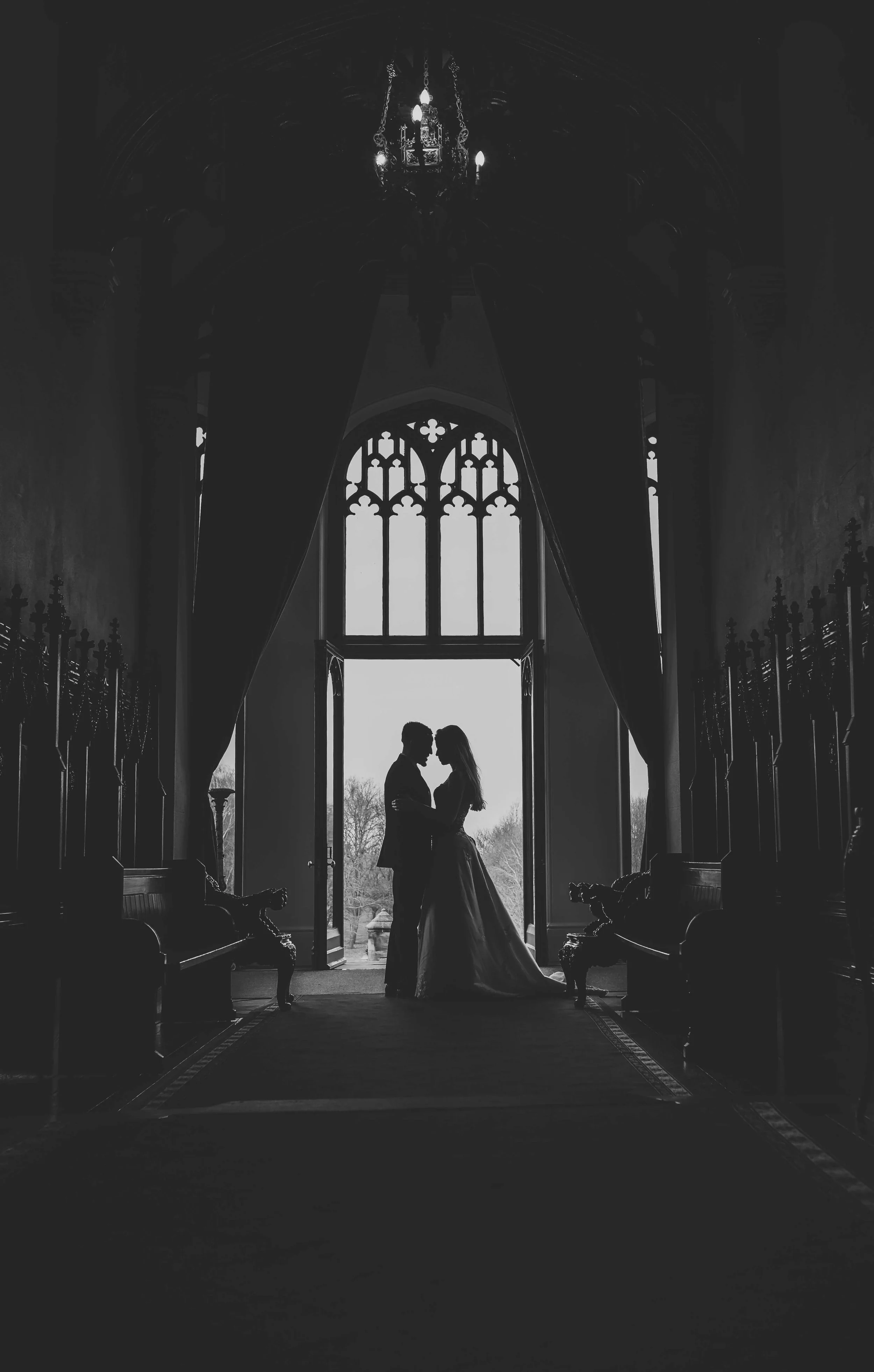 Silhouetted couple standing close together in front of a large open window with Gothic-style stained-glass designs, inside a historic building with ornate decor and a chandelier hanging from the ceiling.