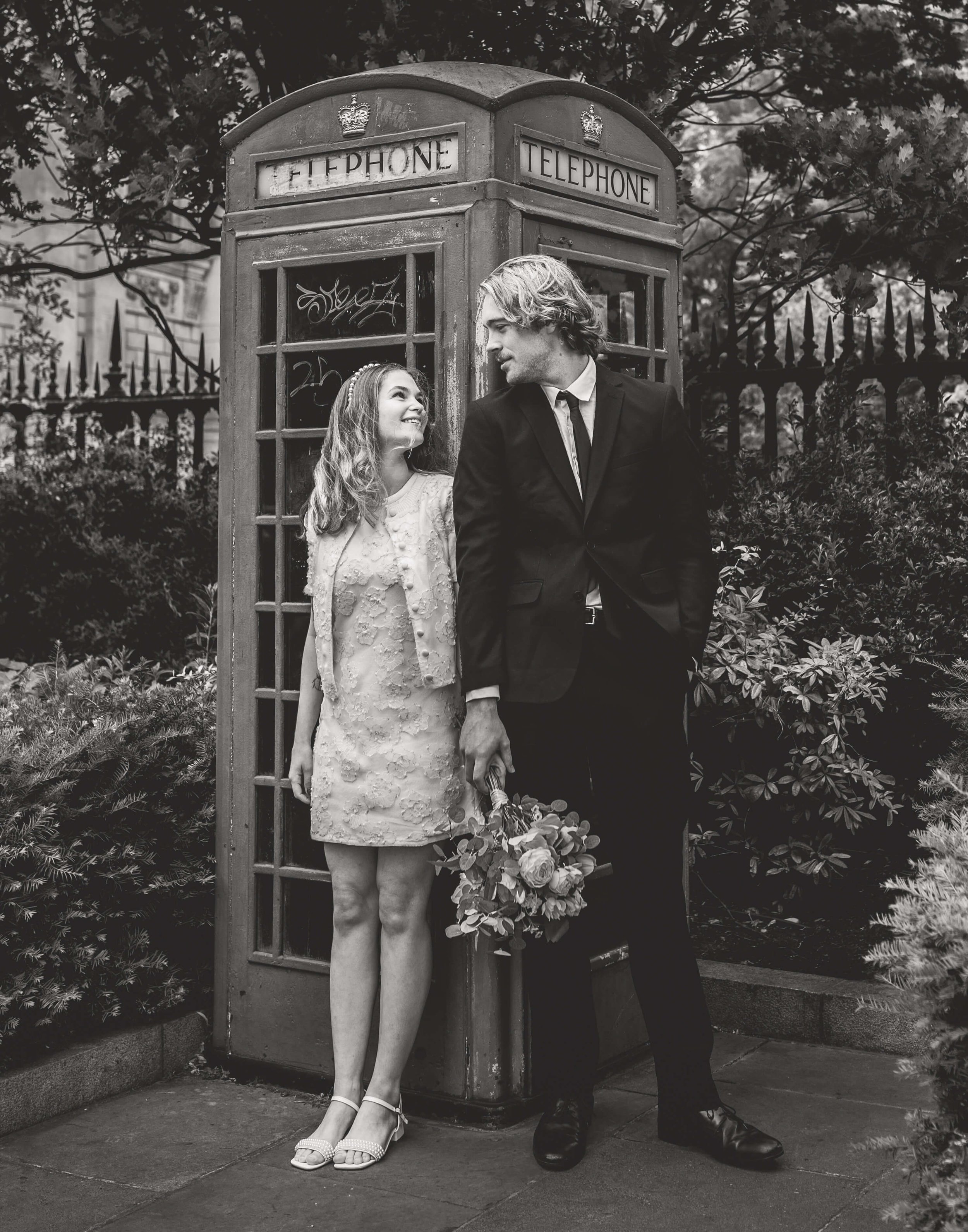 A black and white photo of a young woman and a man standing next to a vintage telephone booth outdoors. The woman, dressed in a light-colored dress and heels, looks happily at the man, who is holding a bouquet of flowers and dressed in a dark suit, w