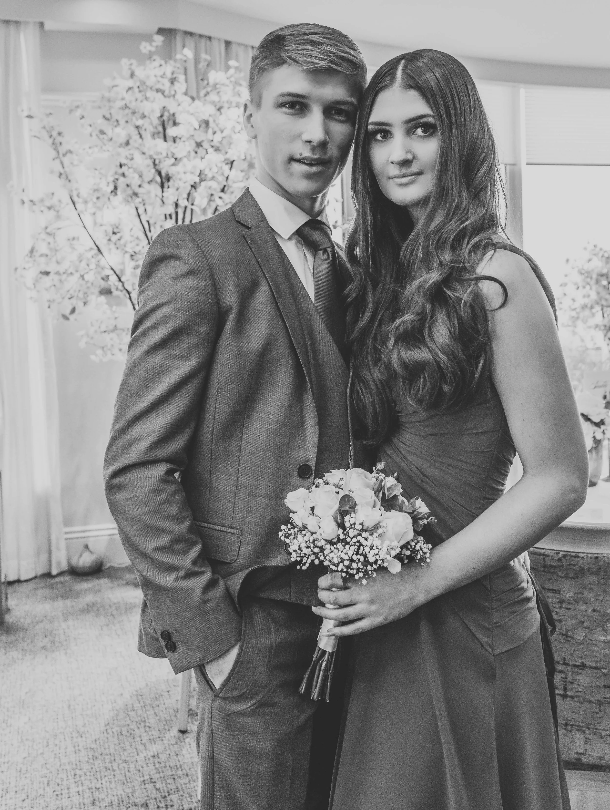 A young man and woman dressed in formal attire, standing indoors, with the woman holding a bouquet of flowers, both looking at the camera.