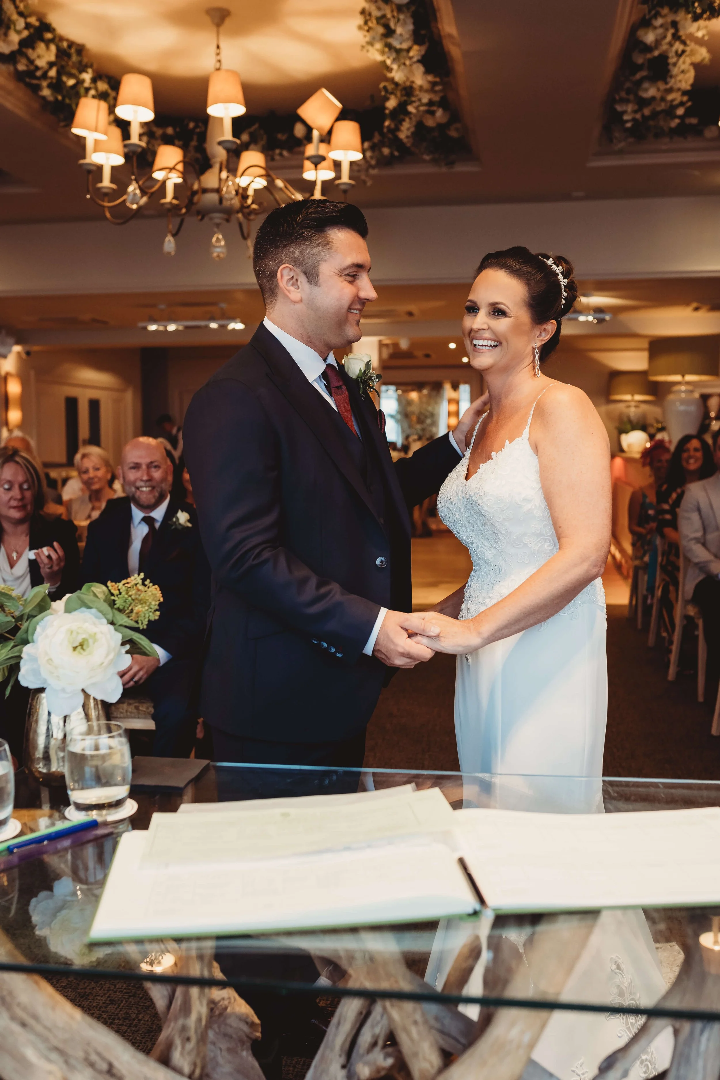 A bride and groom having a wedding ceremony indoors, holding hands and smiling at each other, with guests seated in the background.