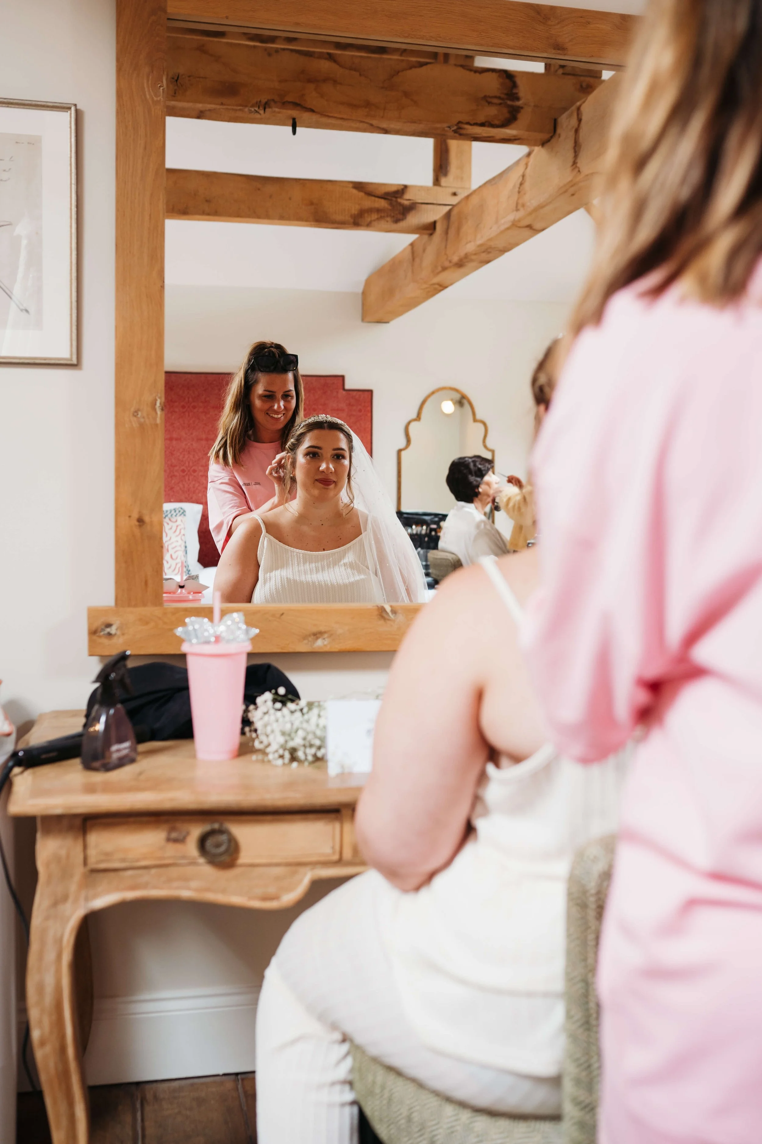 Brunette bride sitting at a vanity table in a dressing room, with a makeup artist applying makeup behind her. The scene is reflected in a mirror.