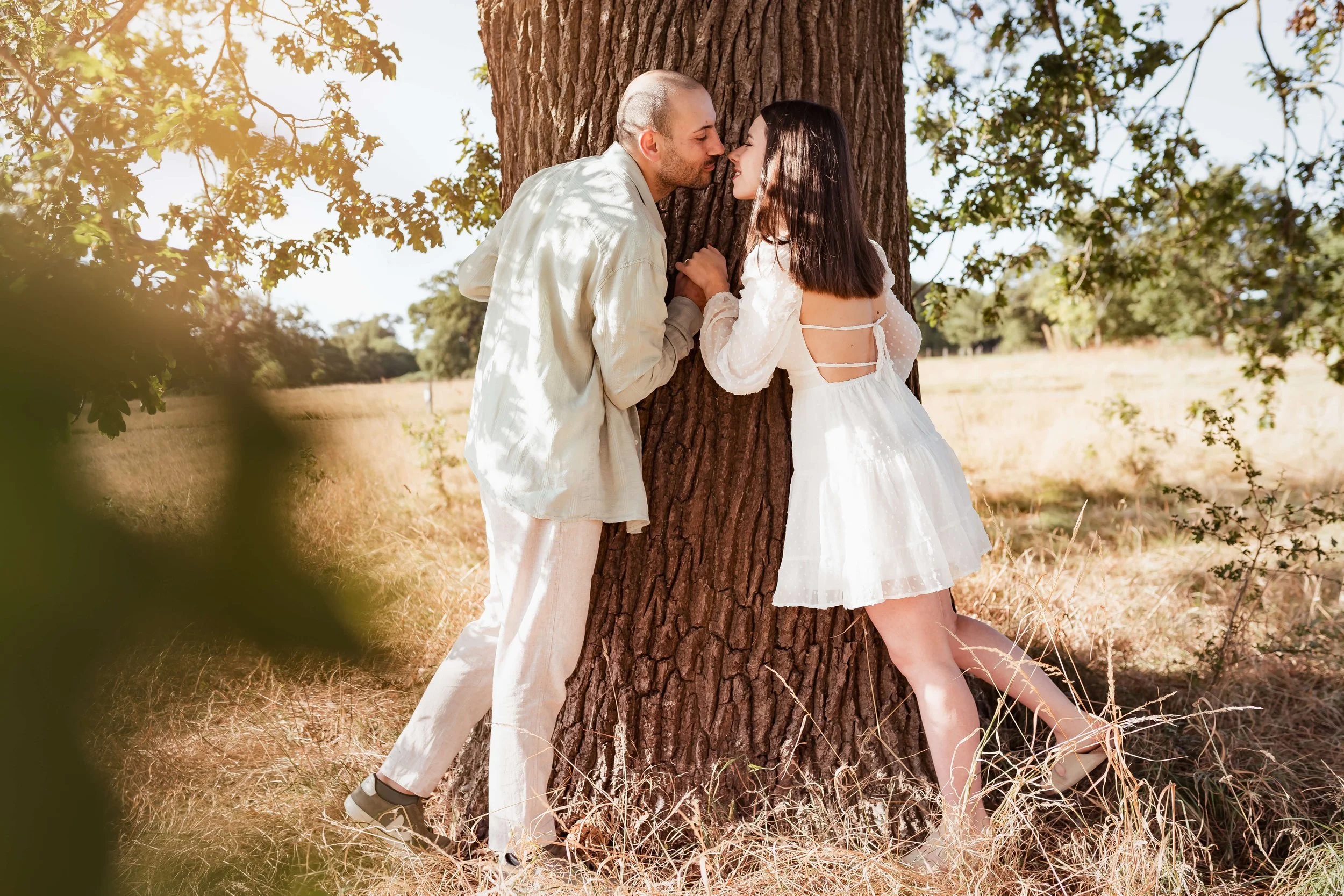A couple is leaning against a tree in a sunny, open field, about to kiss.