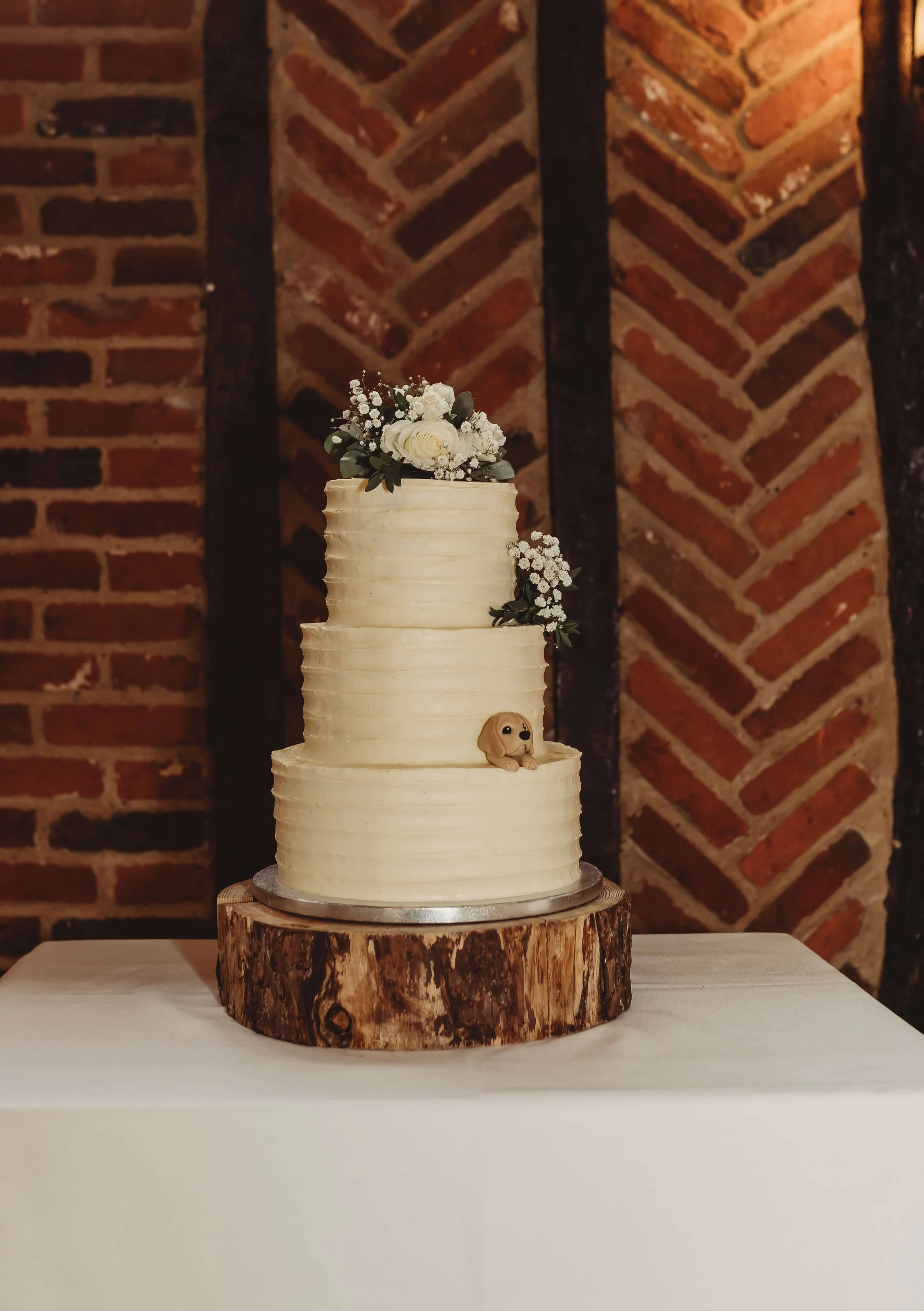 Three-tier wedding cake with white frosting, decorated with white flowers, greenery, and a small dog figurine, sitting on a wooden cake stand against a brick wall background.