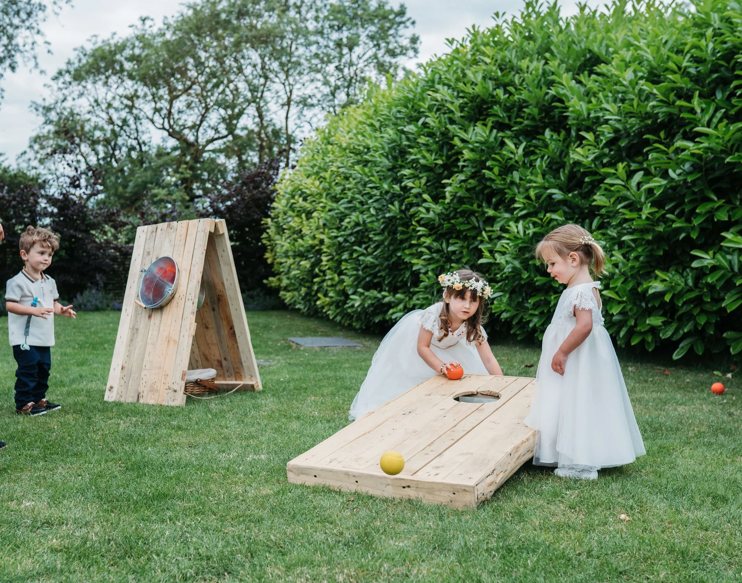 Four children playing croquet on a lawn with green bushes and trees in the background. Two girls in white dresses with floral headbands are focused on the game, one girl is holding a croquet ball, and a boy in a gray shirt and navy pants stands nearb