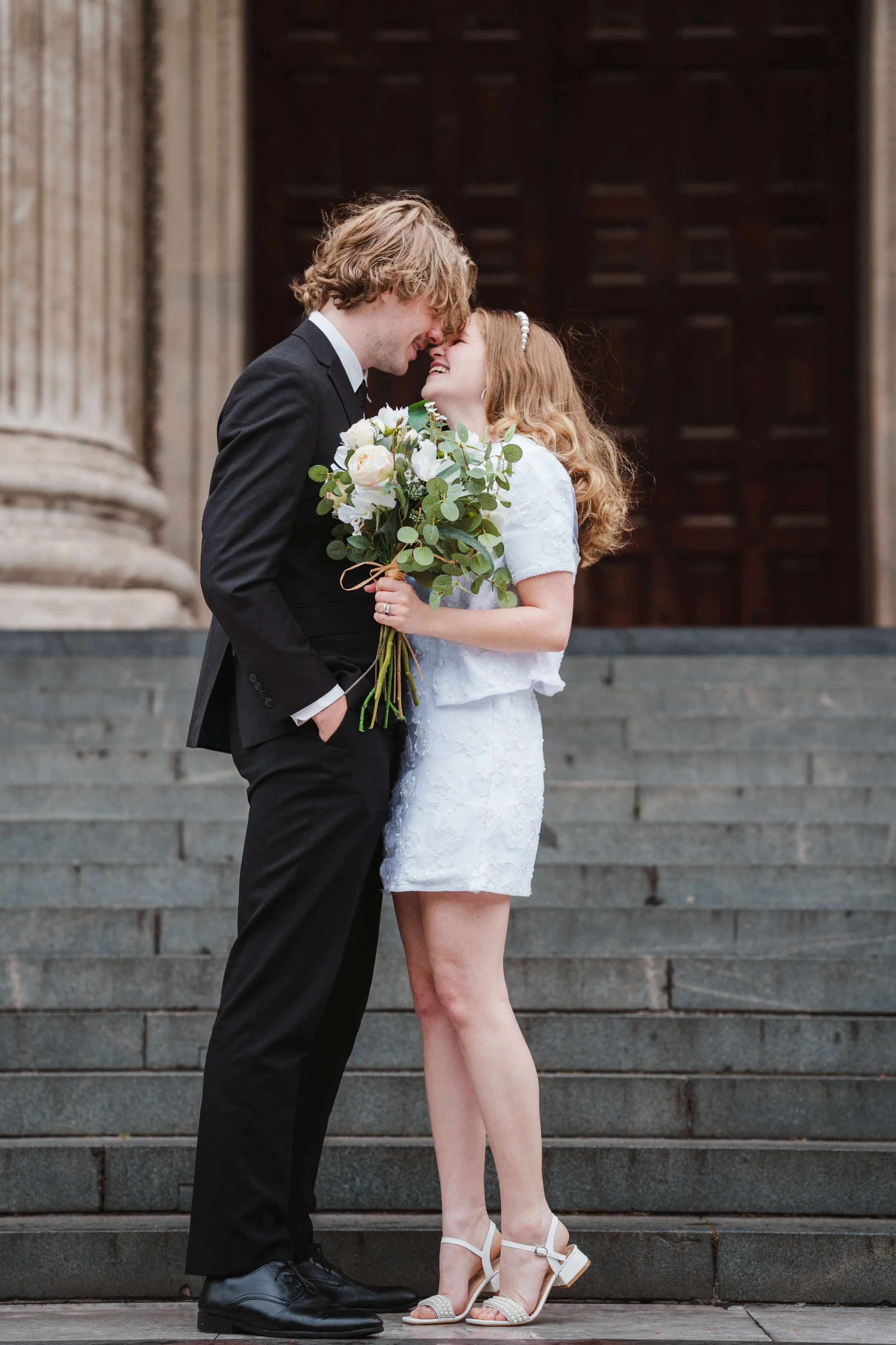 A couple in wedding attire standing close together on steps, smiling, with their foreheads touching. The woman is holding a bouquet of white flowers and greenery. The man is wearing a black suit and the woman a white dress with heels.