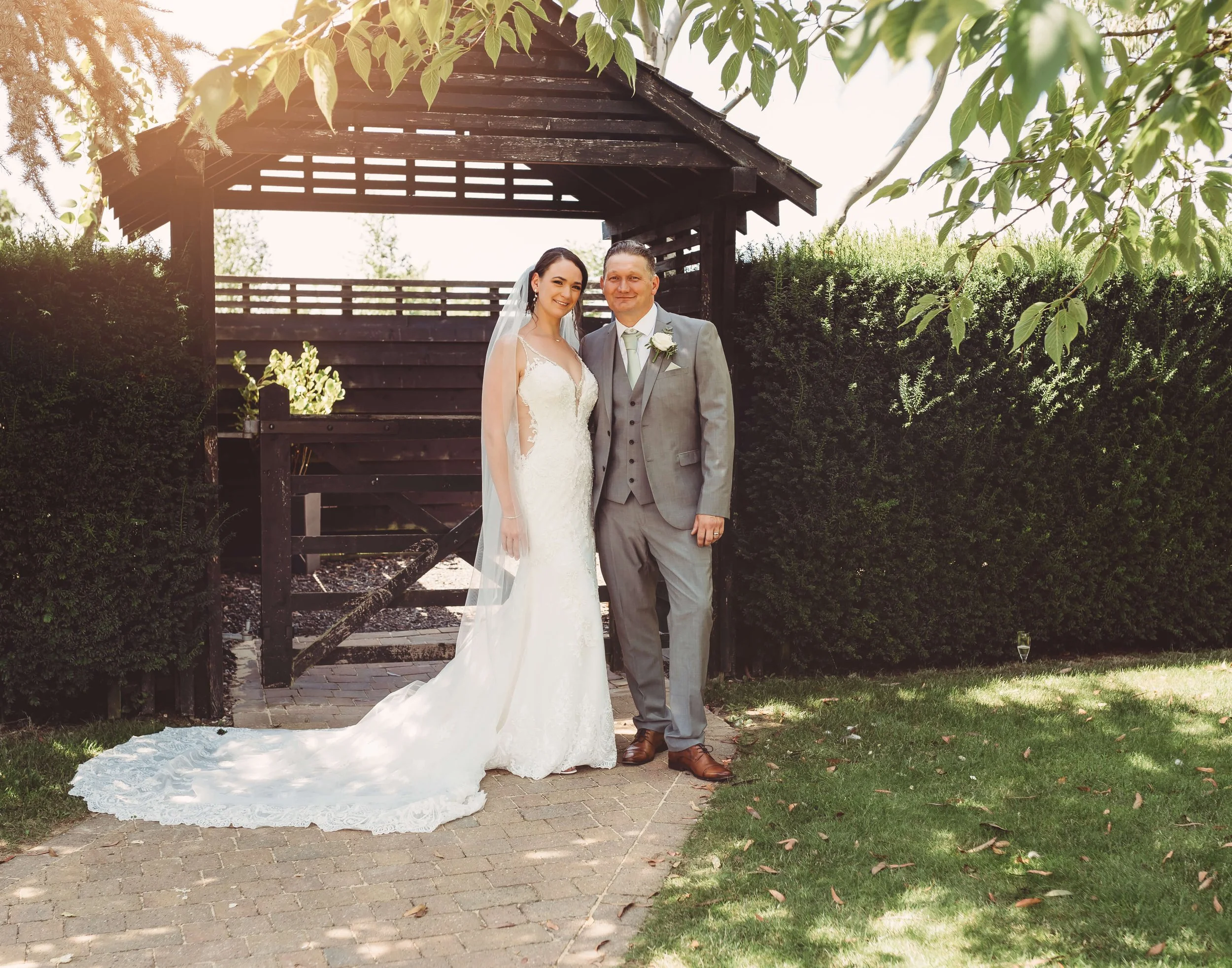 A bride and groom standing together outdoors on a sunny day during their wedding, with the bride in a white lace wedding dress and veil, and the groom in a light gray suit with a white tie and boutonniere.