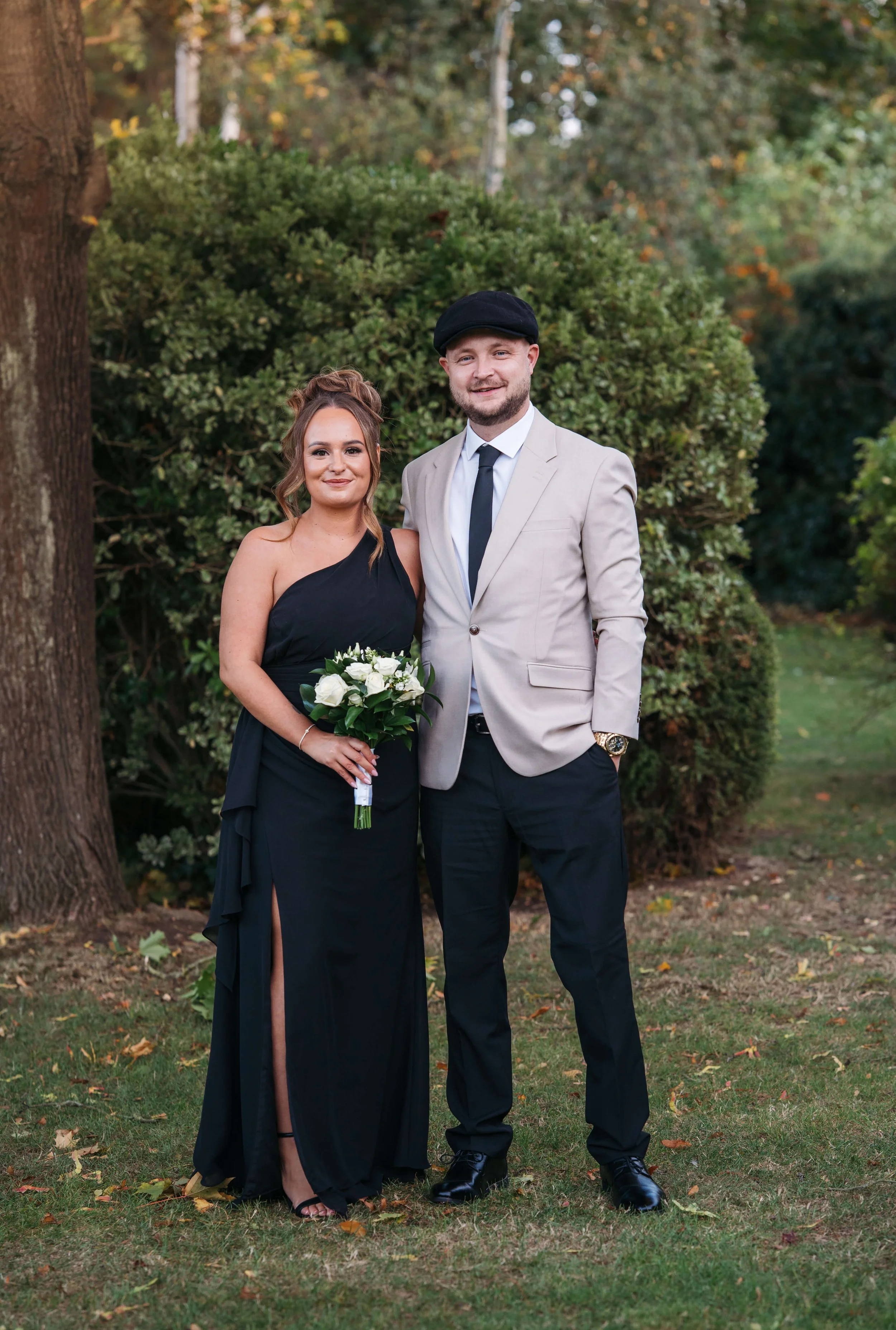 A woman in a black dress holding a bouquet of white roses standing next to a man in a beige blazer, black pants, and a black hat outdoors in a park with trees and greenery.