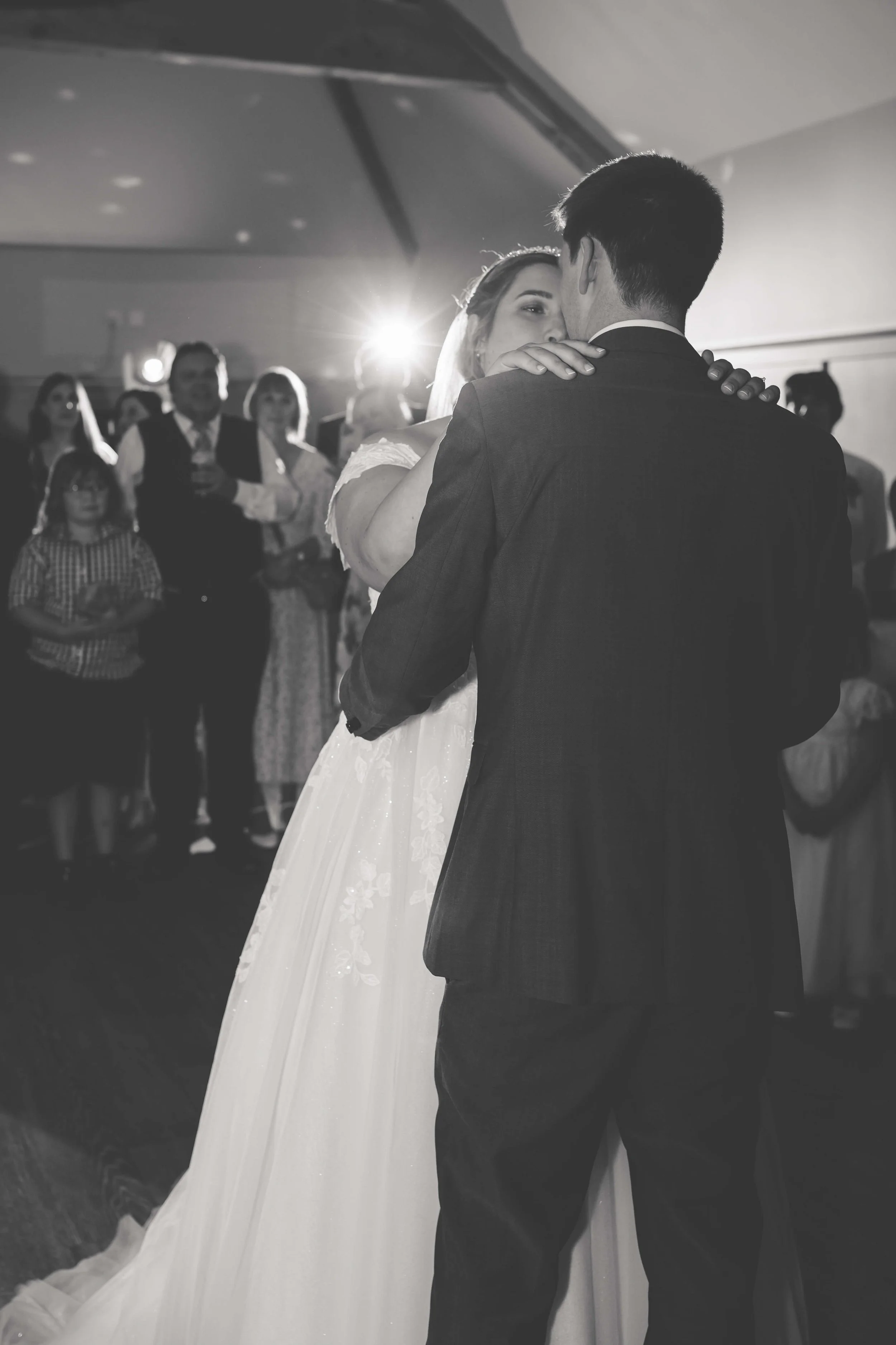 A bride and groom share their first dance at a wedding reception, surrounded by guests, in black and white photo.