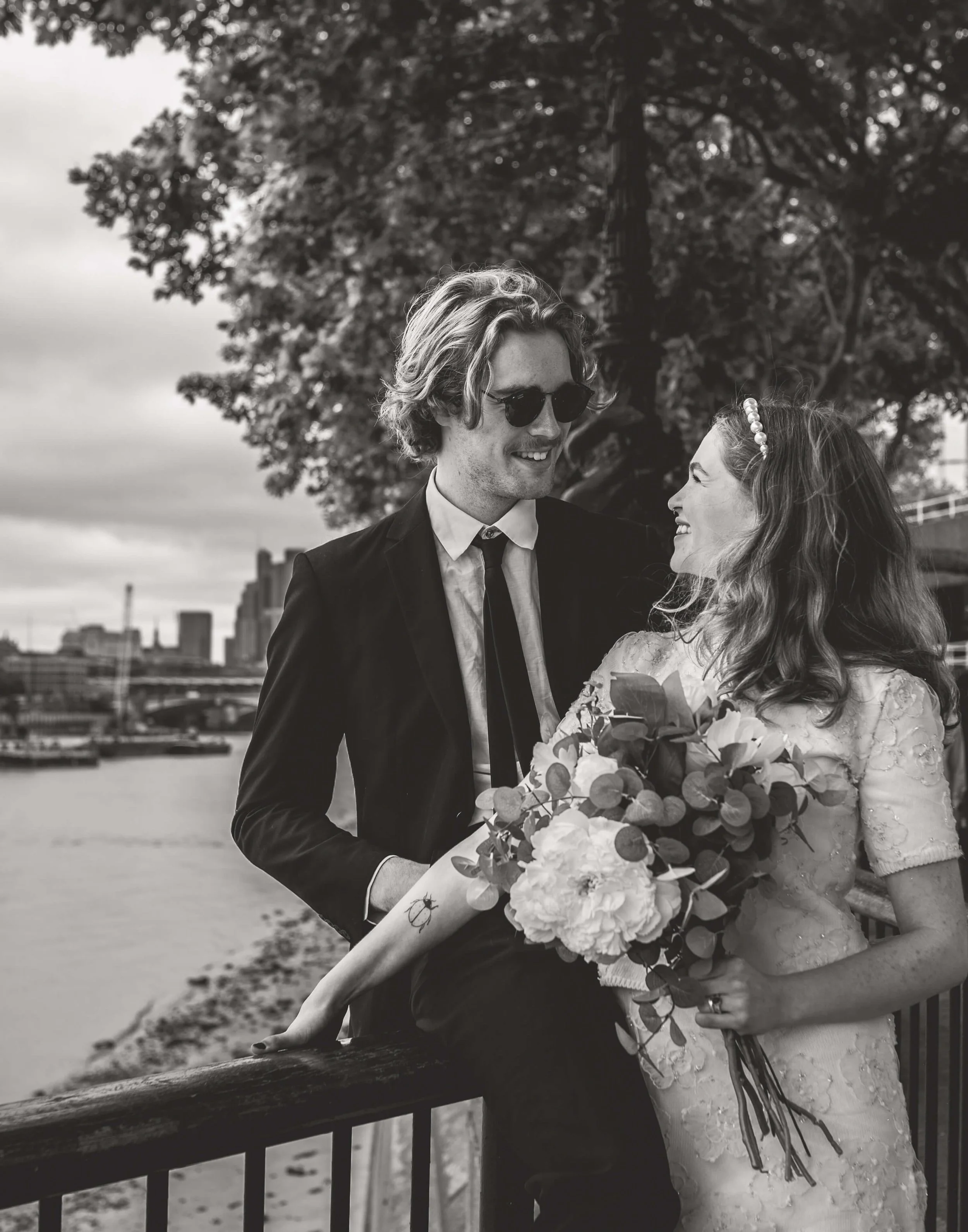 A black and white photograph of a couple at a riverside. The man is wearing a suit, tie, and sunglasses, and the woman is in a wedding dress holding a bouquet of flowers. They are smiling and looking at each other, standing close next to a fence with