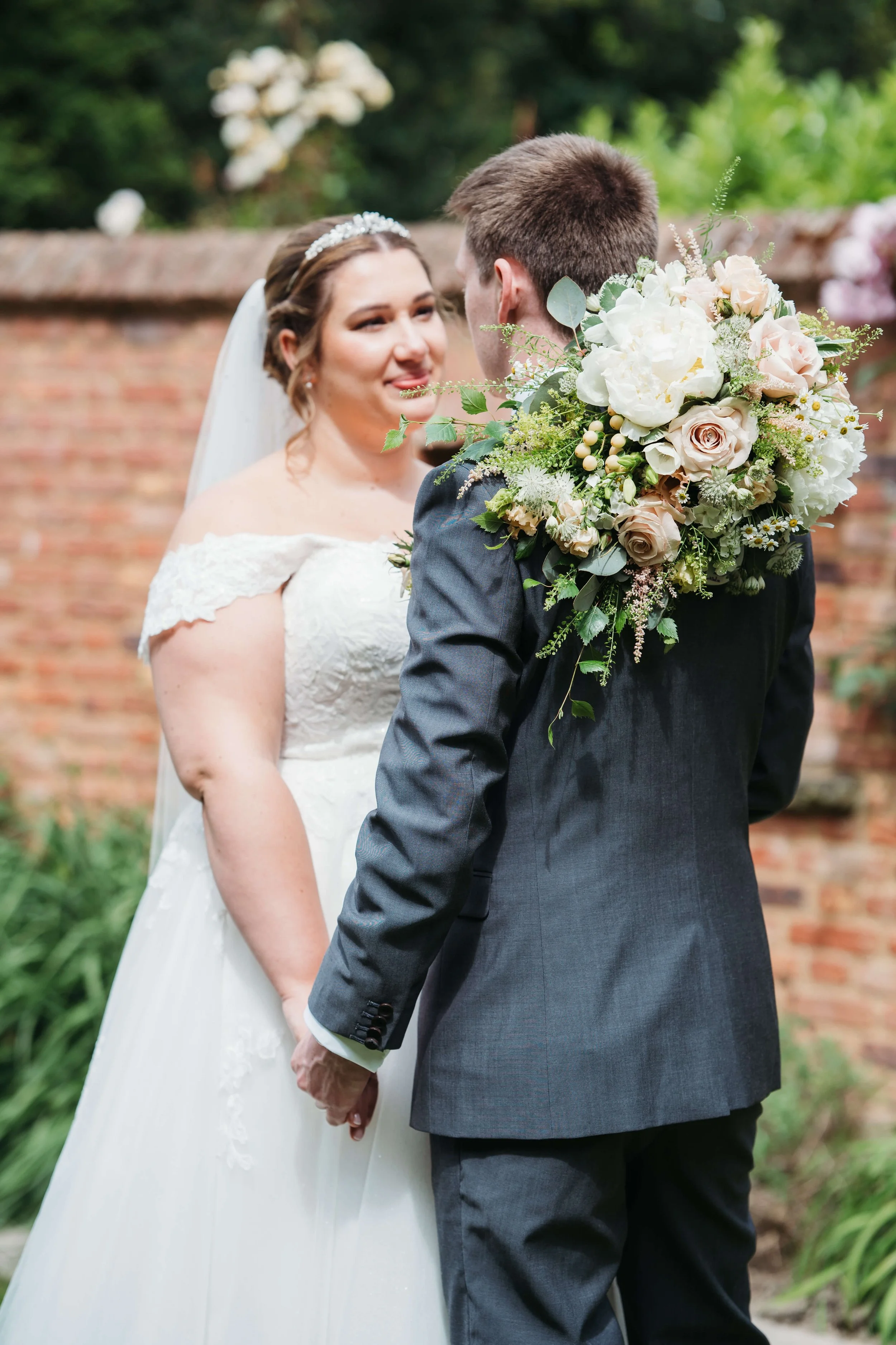 A bride and groom facing each other in an outdoor wedding ceremony. The bride wears a white lace off-shoulder wedding gown and a tiara, while the groom wears a dark suit and holds a large bouquet of white and blush roses, greenery, and berries.