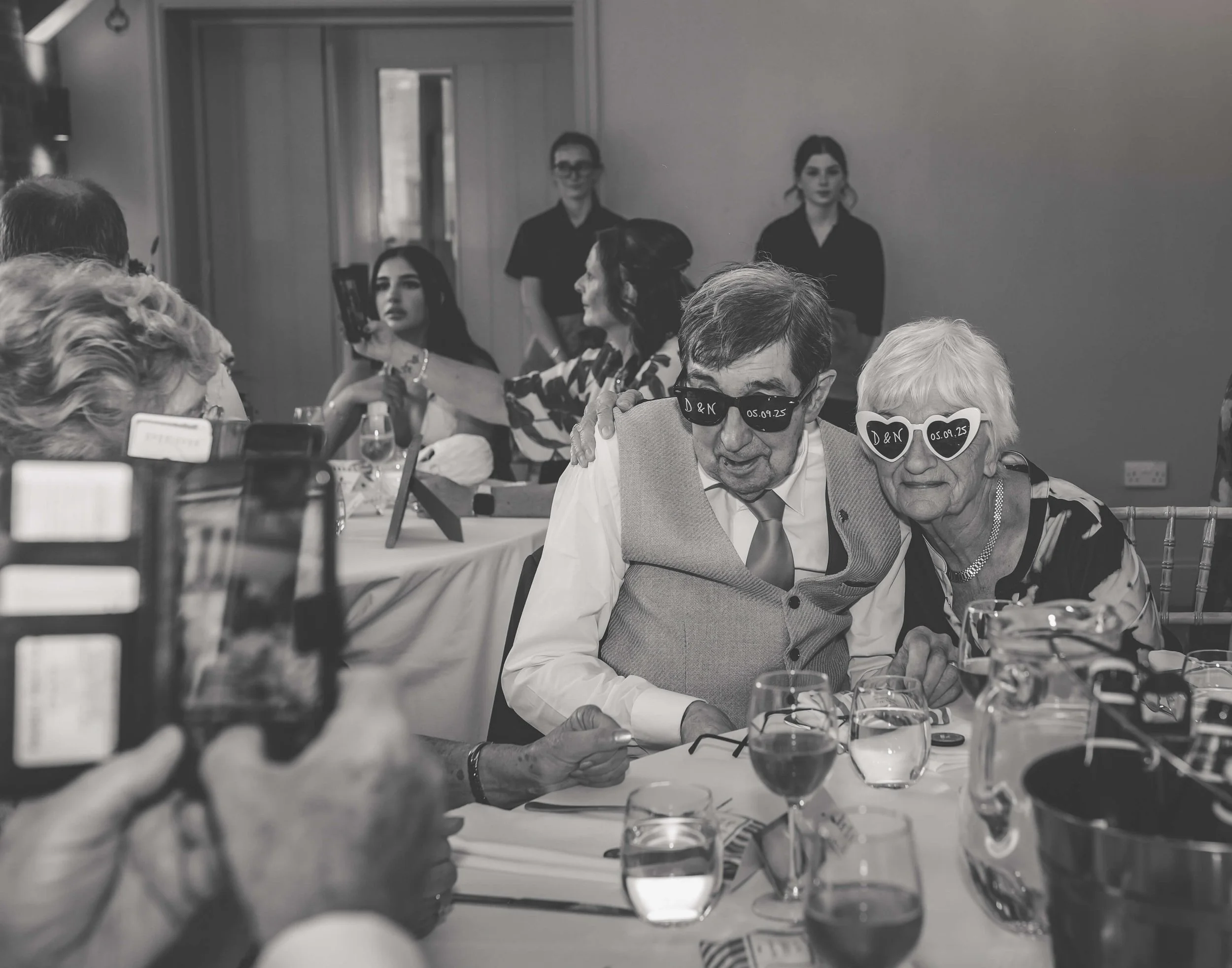 Older man and woman at a celebration or party, both wearing sunglasses with inscriptions, sitting close together at a table with drinks, with other guests and staff in the background.