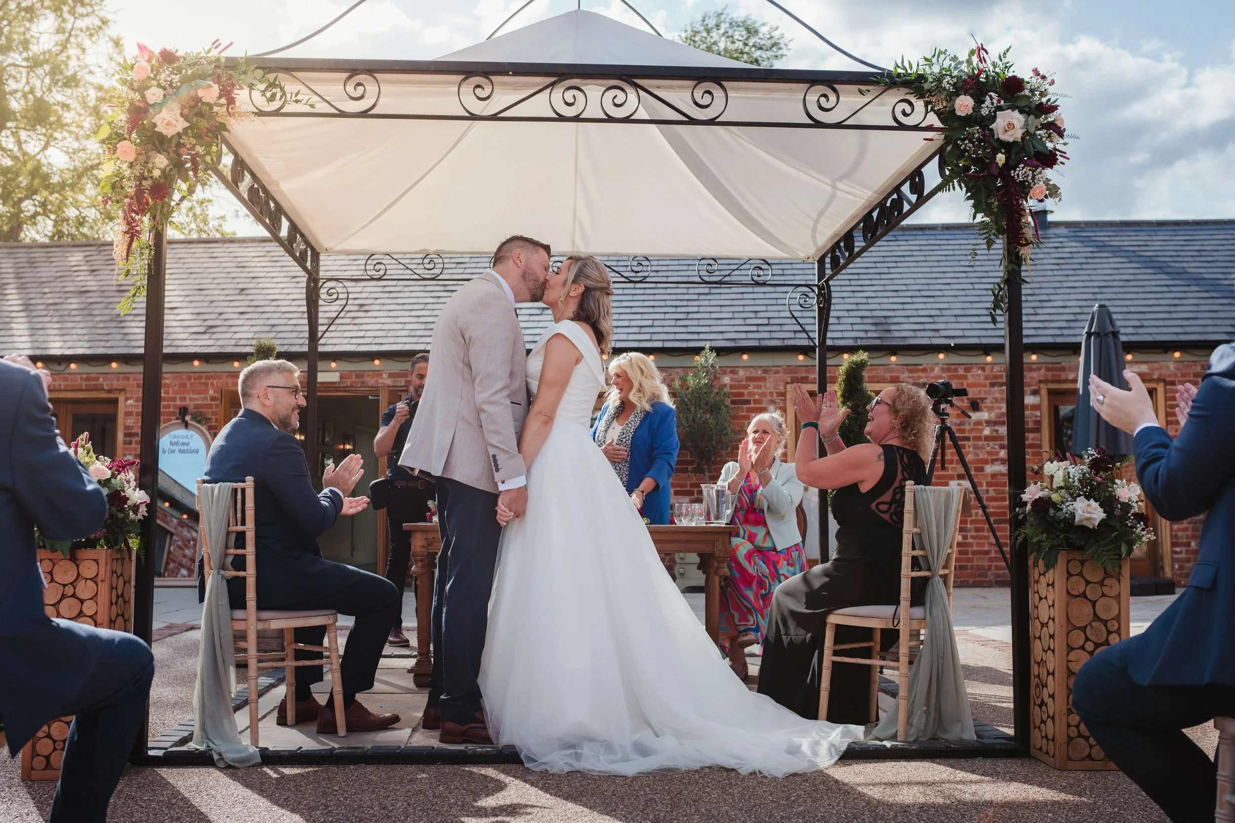 A couple shares a kiss during their wedding ceremony under a decorated outdoor canopy, surrounded by seated guests and sunlight.