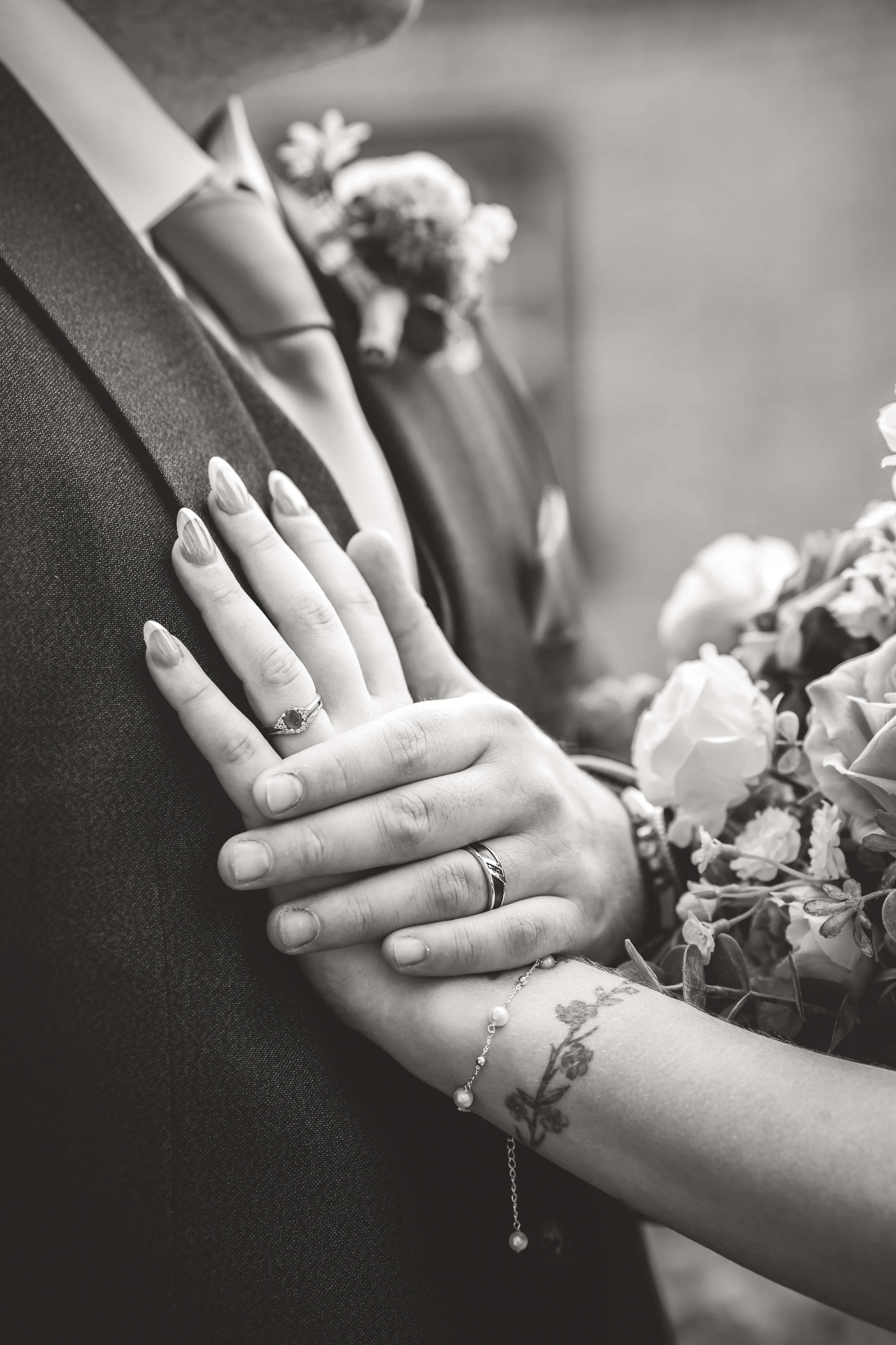 Close-up of a couple’s hands, showing wedding rings and a floral bracelet, with a bouquet of flowers and a groom in a suit in the background, black and white photo.