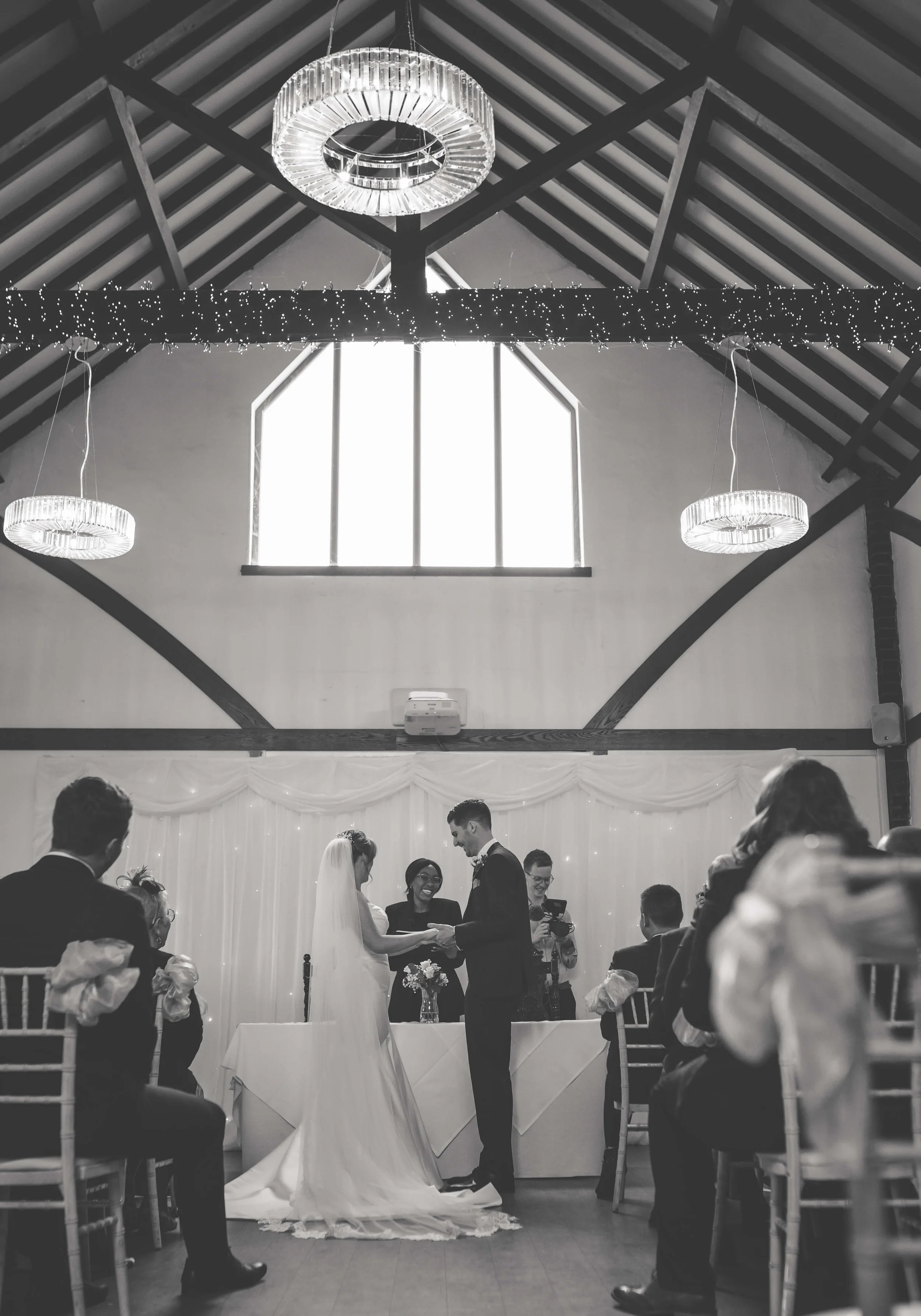 A black and white photograph of a wedding ceremony inside a decorated venue. The bride and groom are exchanging rings at the altar, surrounded by seated guests, with the bride wearing a veil and gown, and the groom in a suit. The background features 