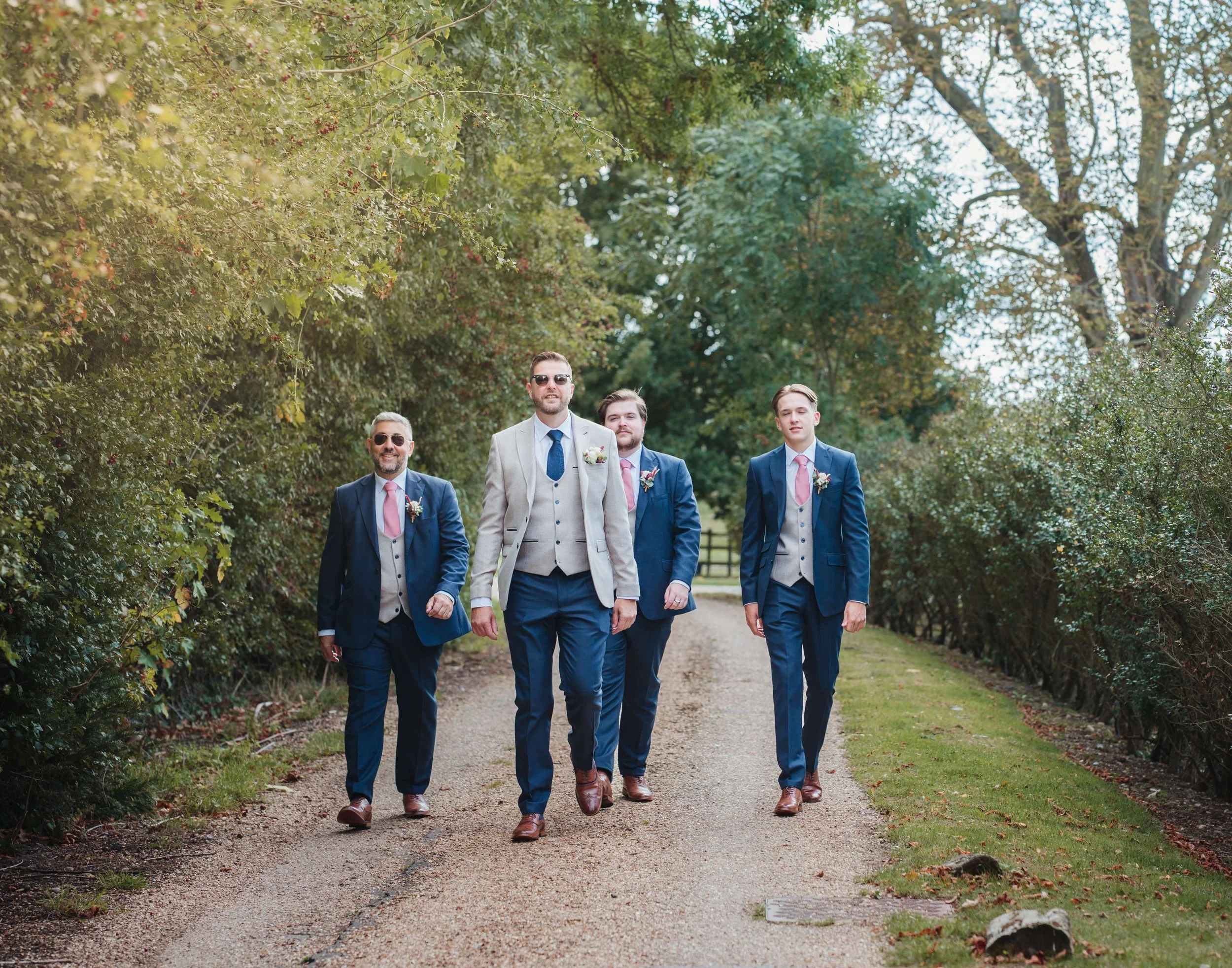 Five men walking on a gravel path surrounded by trees, dressed in formal suits and ties, with boutonnieres, in a park setting.
