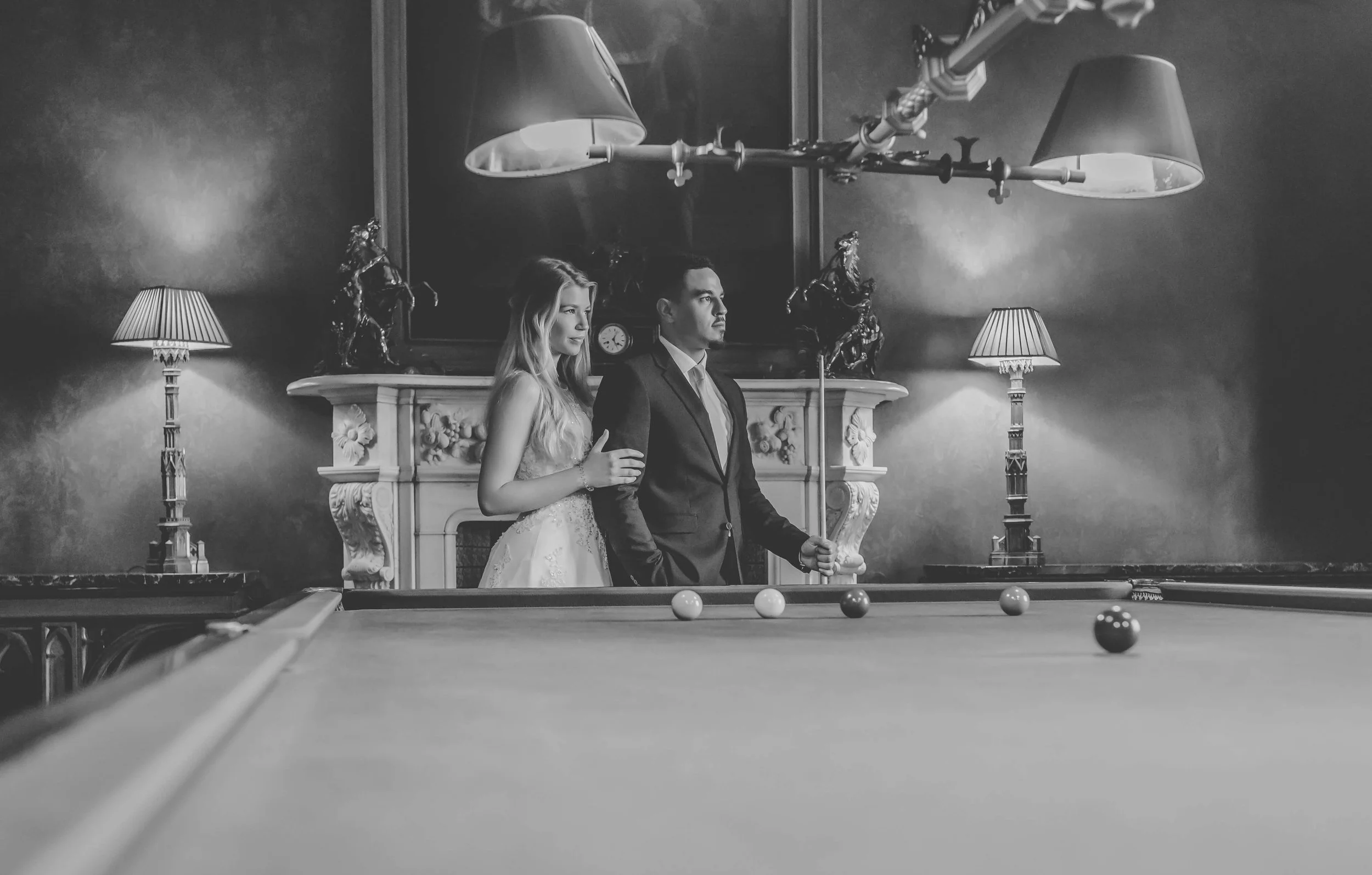 A black-and-white photo of a bride and groom standing next to each other in a luxurious room. The bride is in a white wedding dress, and the groom is in a dark suit, holding a cue stick. They are beside a pool table, with balls set up for a game. The