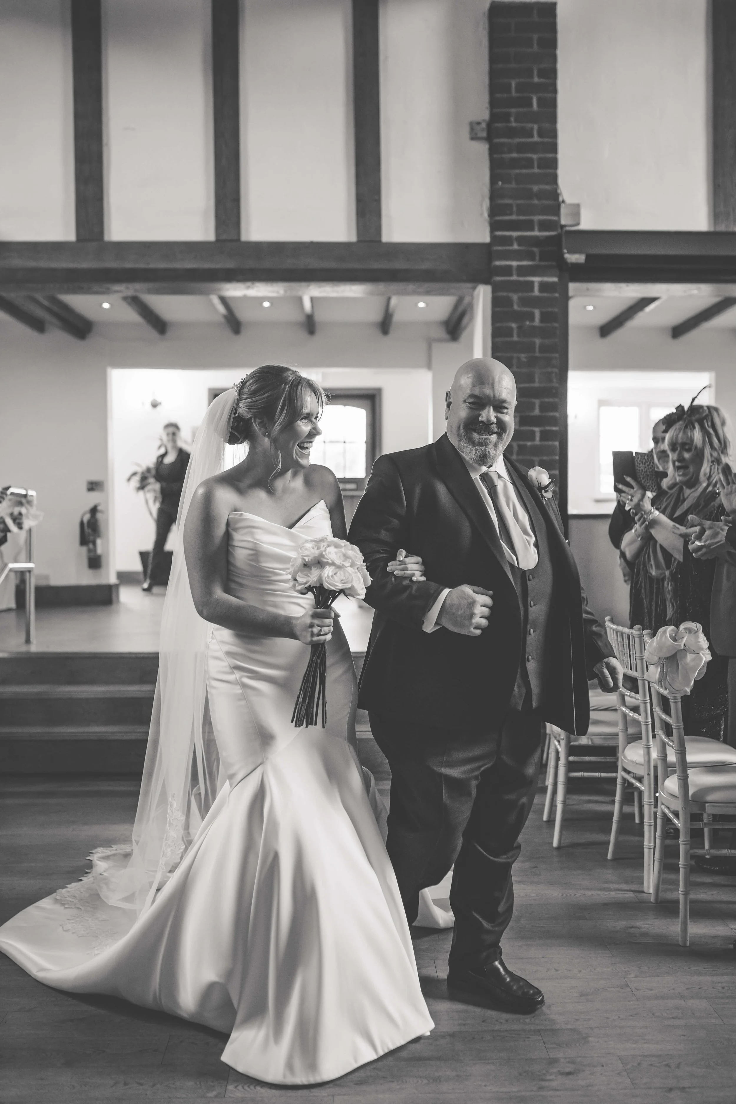 Black and white photo of a bride in a wedding dress walking down the aisle, being escorted by a man, at a wedding reception with guests taking pictures.