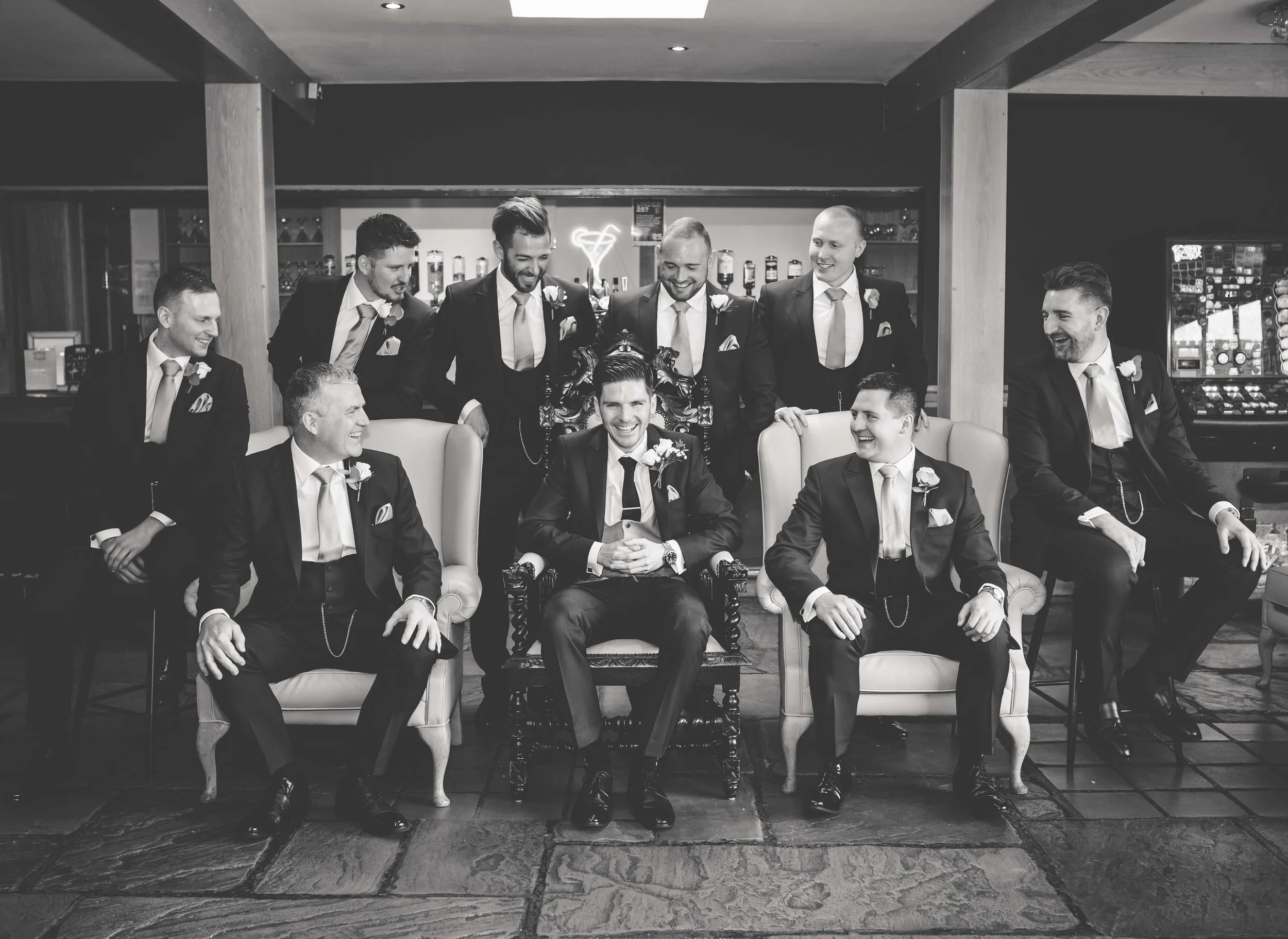 Group of men in suits, some sitting on chairs and some standing behind, smiling and enjoying themselves in a bar or lounge setting.