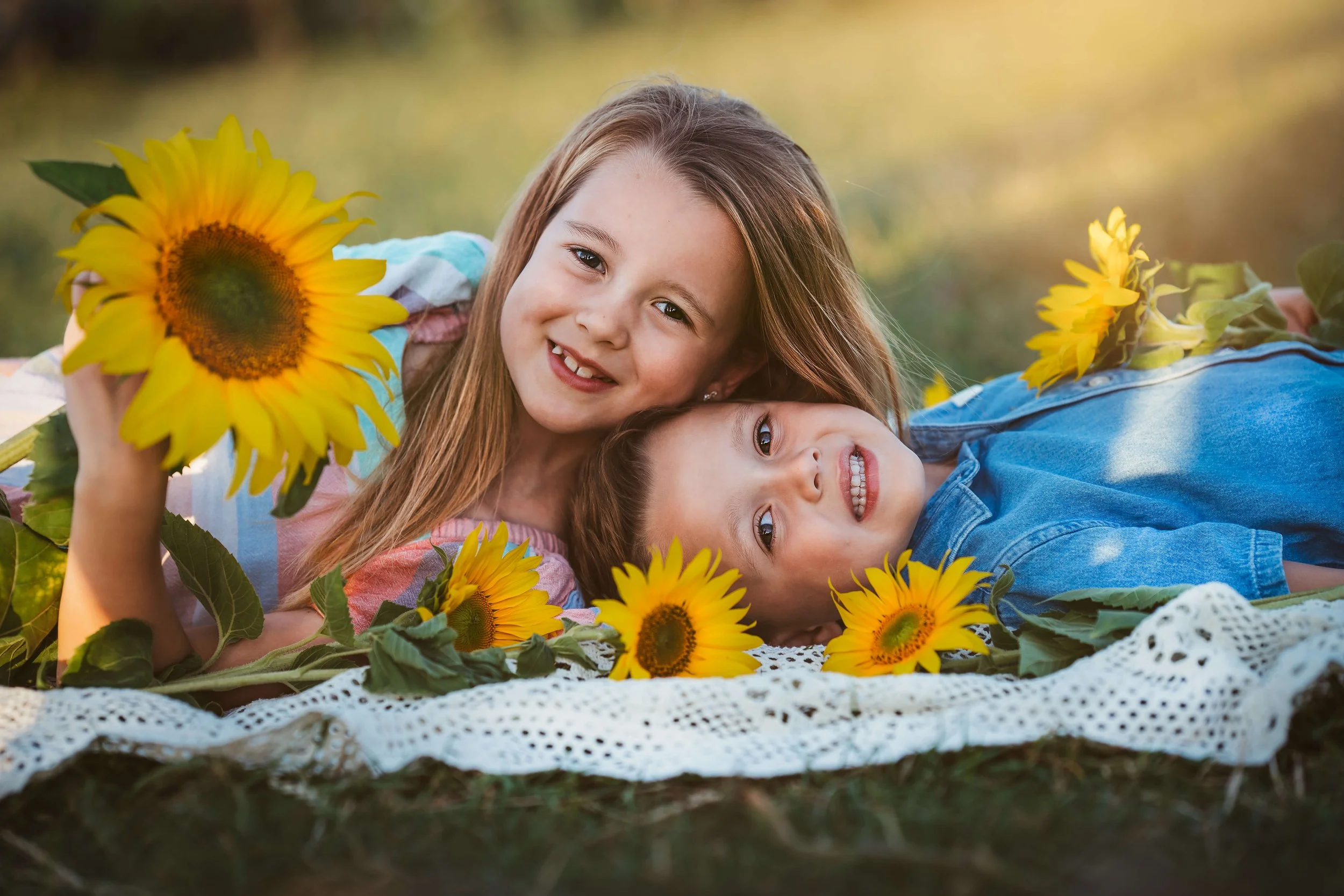 Two children, a girl and a boy, lying on a white blanket surrounded by sunflowers in an outdoor setting during daylight, smiling at the camera.