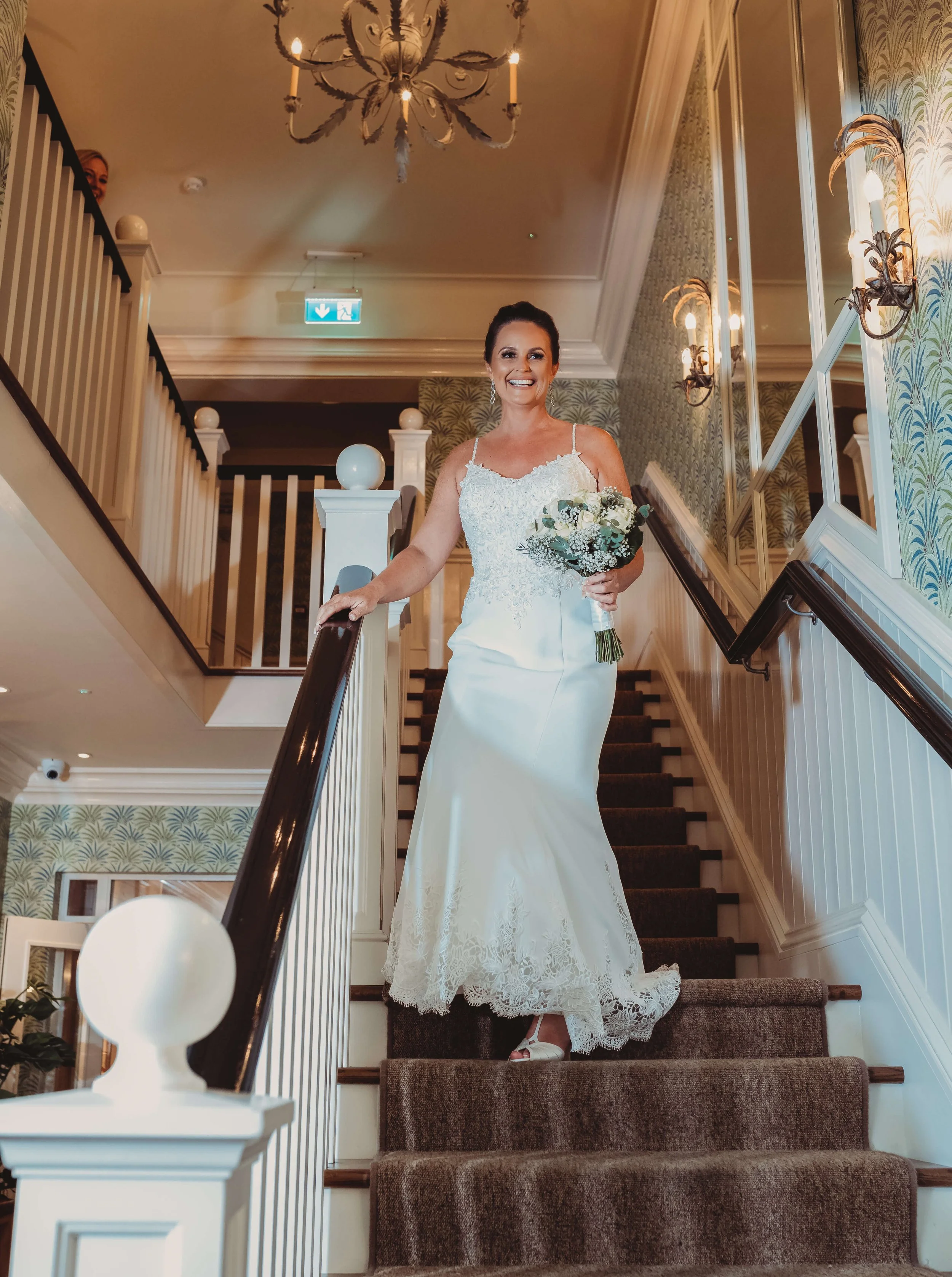 A bride in a white wedding gown holding a bouquet, descending a staircase inside a decorated venue. She is smiling and looking at the camera.