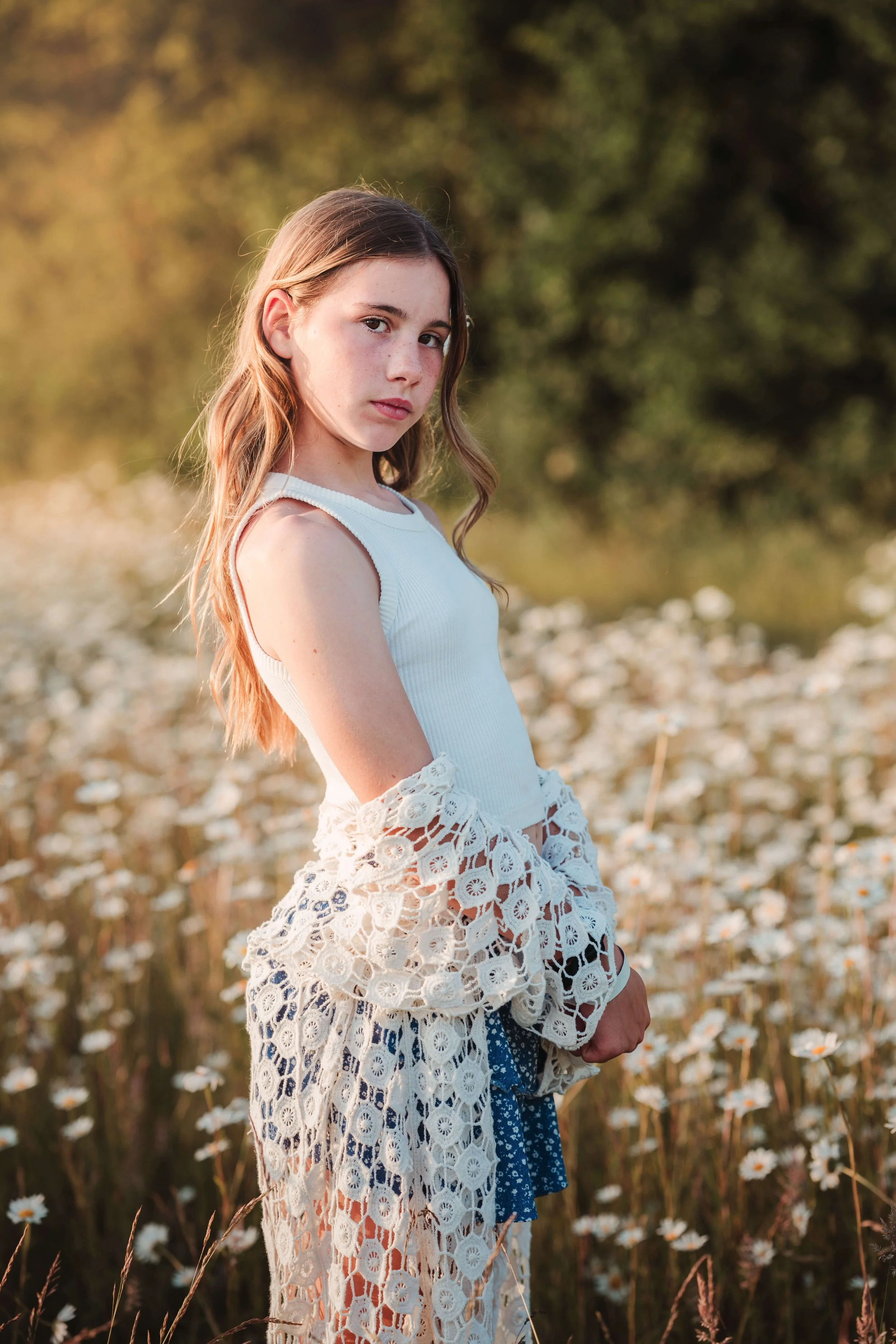 A young girl with long, wavy hair stands in a field of flowers, wearing a sleeveless white top and a white lace jacket, with trees and a blurred background.