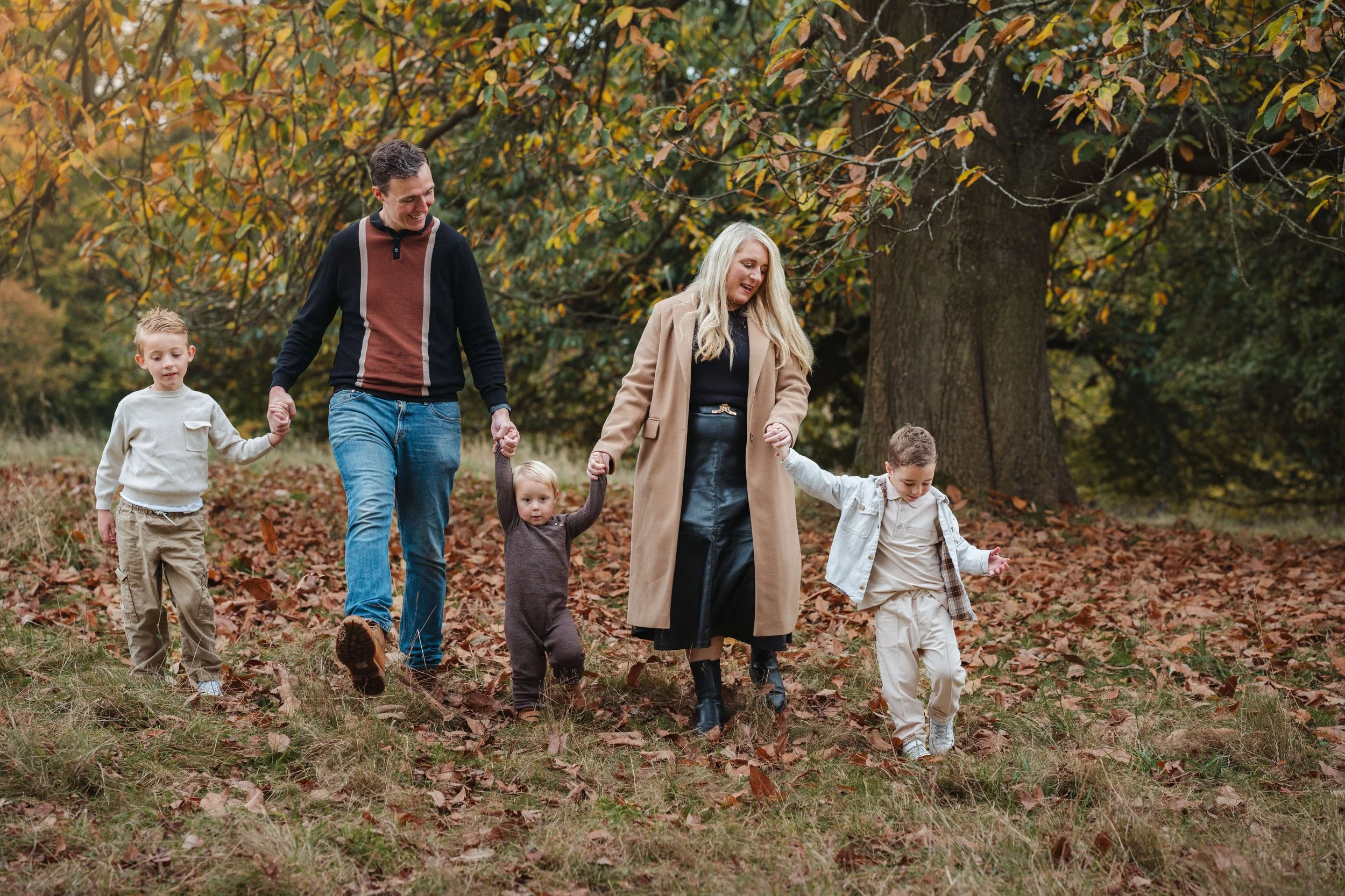 A family of five walking outdoors through fallen leaves in the fall, holding hands and smiling, with trees in the background.