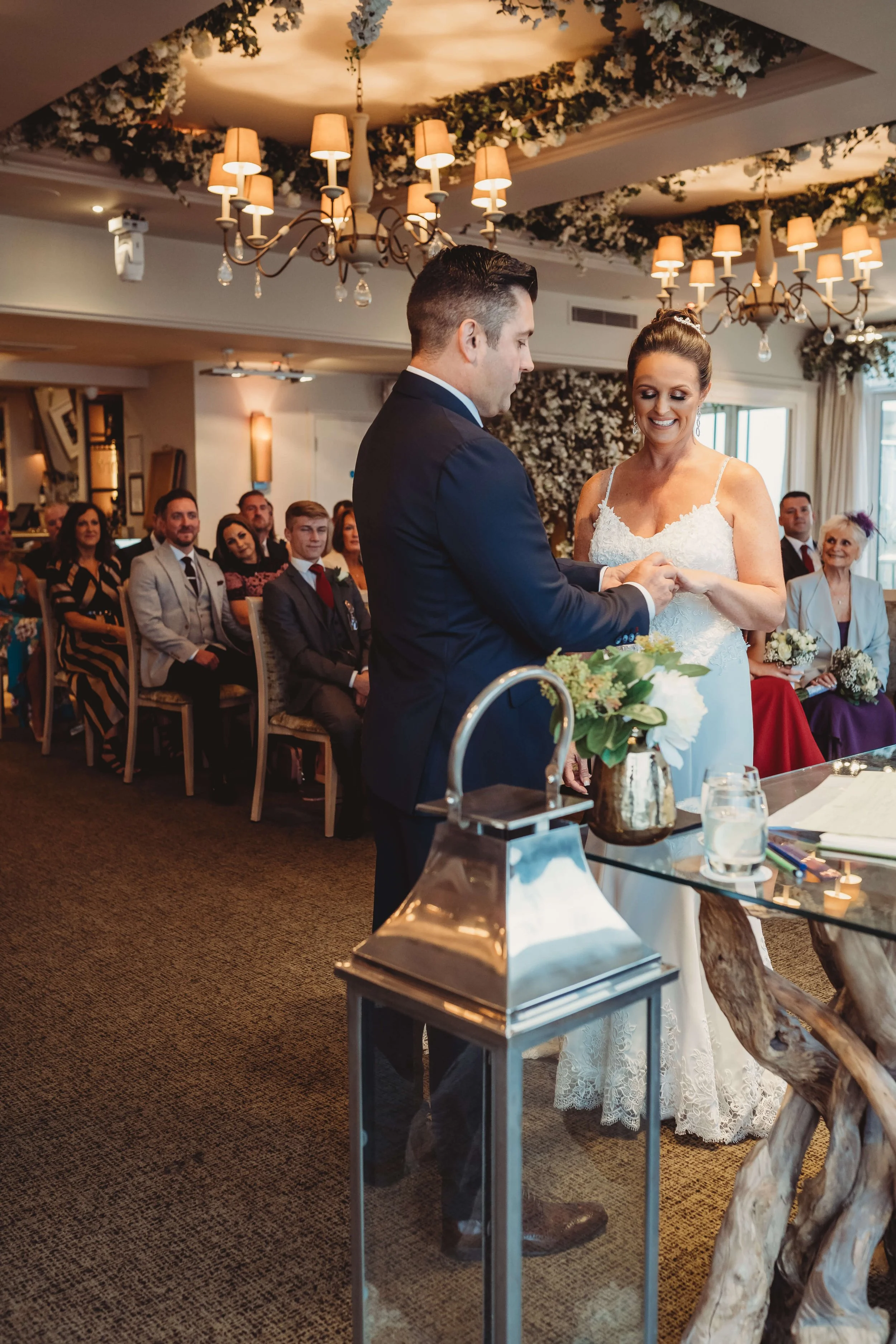A bride and groom exchanging rings during their wedding ceremony in a decorated indoor venue with guests seated in the background.