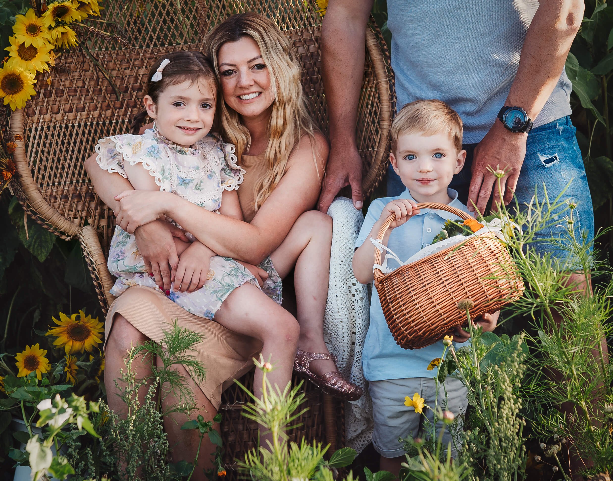 A woman sits on a wicker chair surrounded by two children and flower plants. A girl is sitting on her lap, smiling, and a boy is standing next to her holding a basket with flowers. An adult man stands behind them.