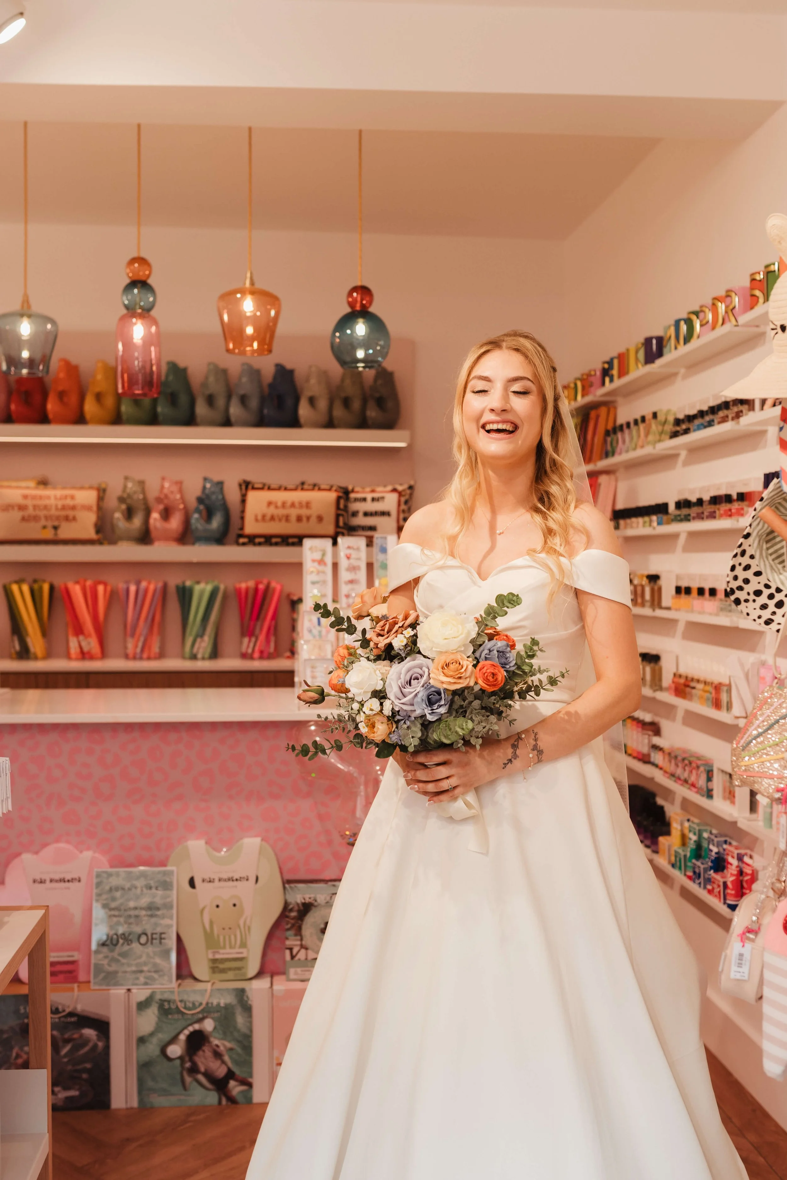 A woman in a wedding dress holding a bouquet of flowers, smiling in a colorful gift shop.
