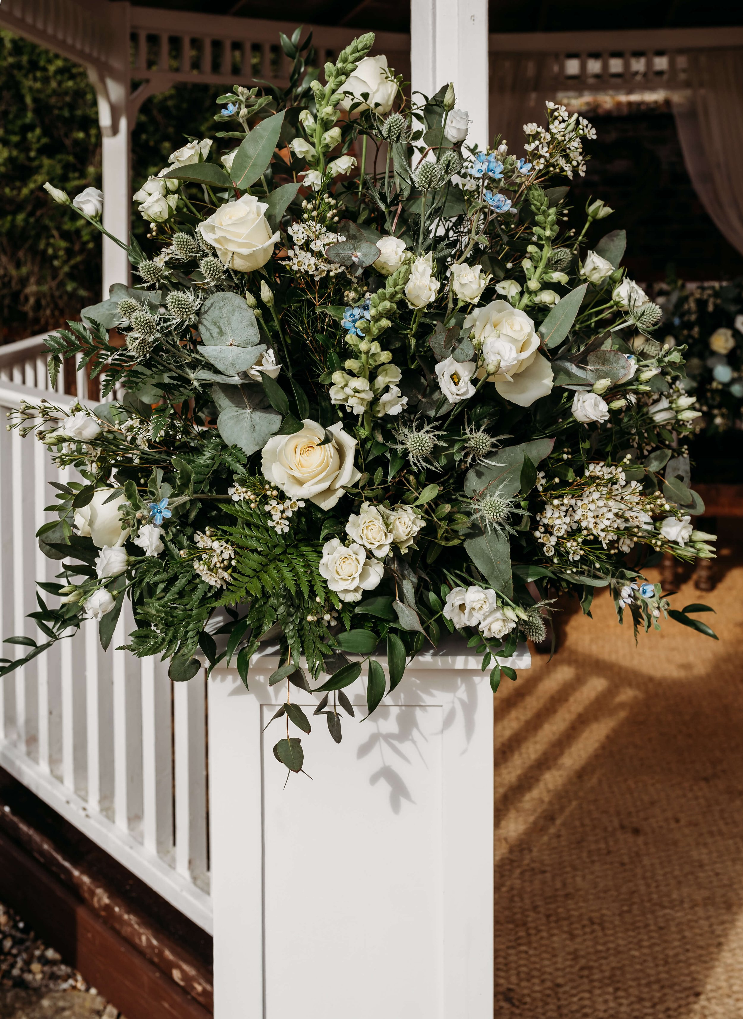 A large floral arrangement with white roses, white lisianthus, small blue flowers, green eucalyptus leaves, and fern, placed in a white container outside a decorated venue.