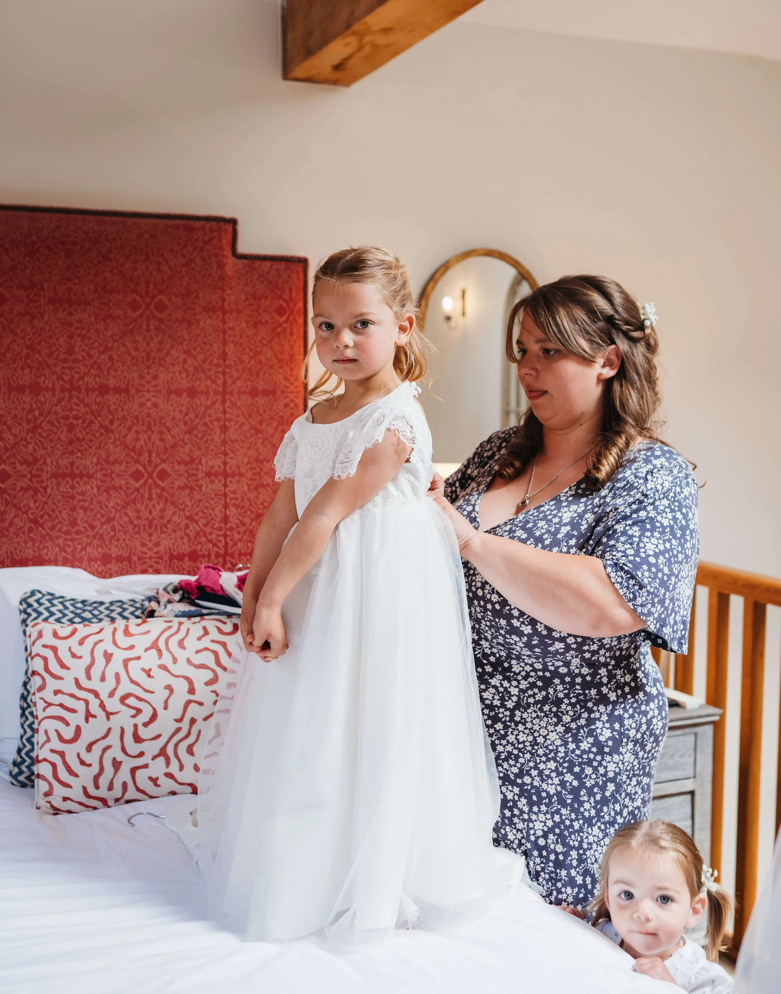 A woman helping a young girl in a white dress onto a bed, with a small girl peeking from behind the bed.