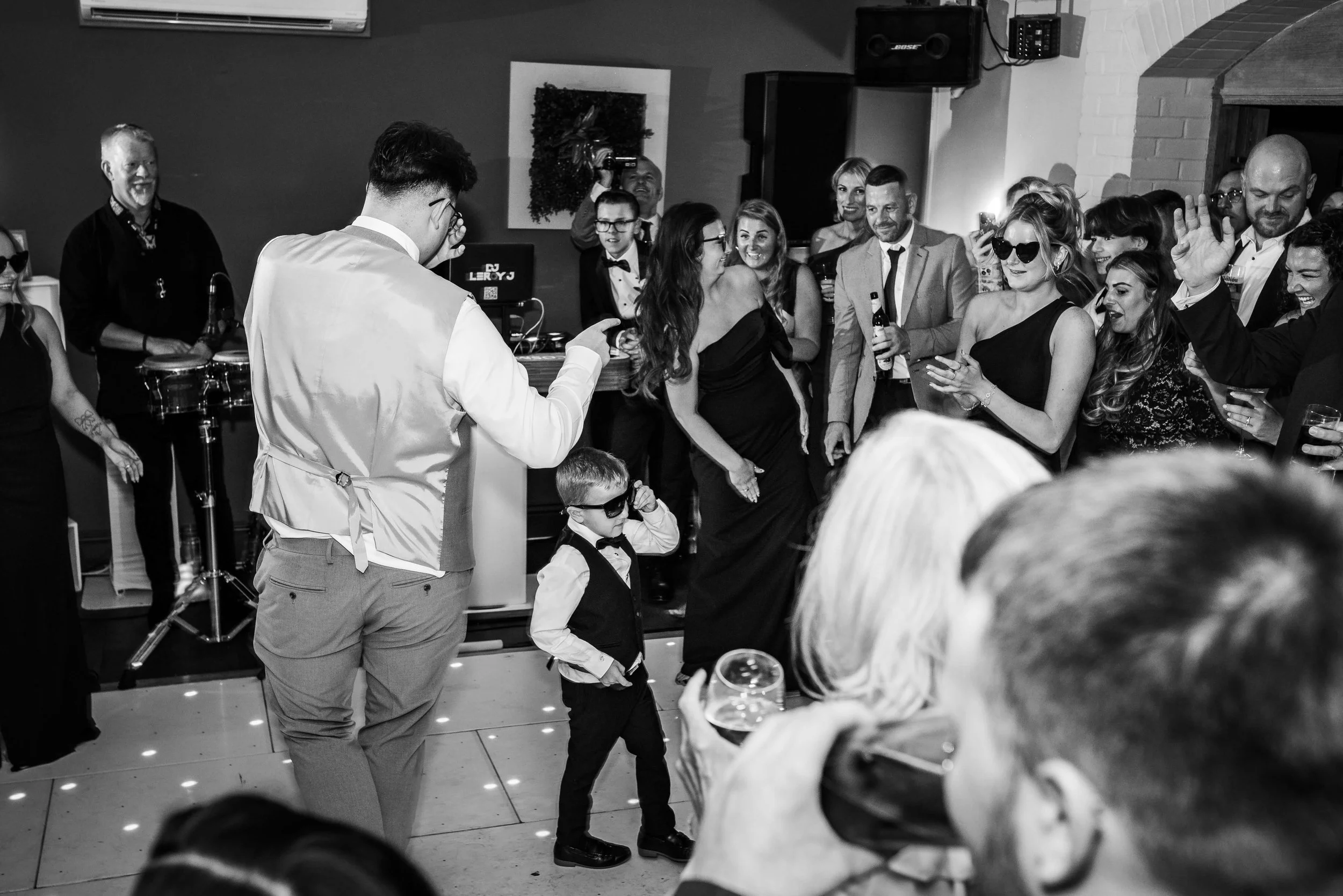 People dancing and celebrating at a party, with a DJ in the background, a young boy wearing sunglasses, and guests holding drinks.