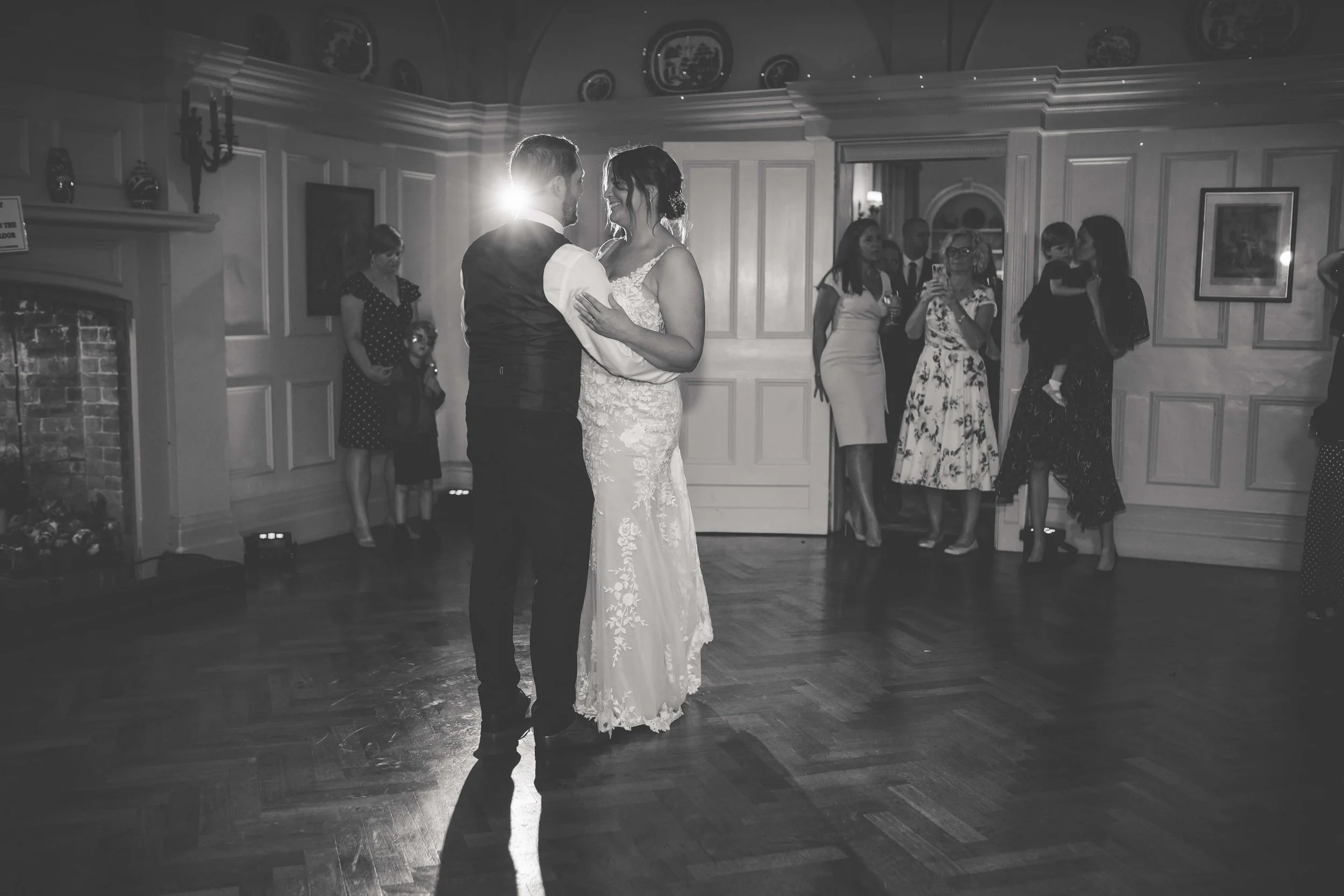 A bride and groom dance in the center of a reception hall with guests watching. The room has wood paneling and framed pictures. The scene is lit with soft lighting.