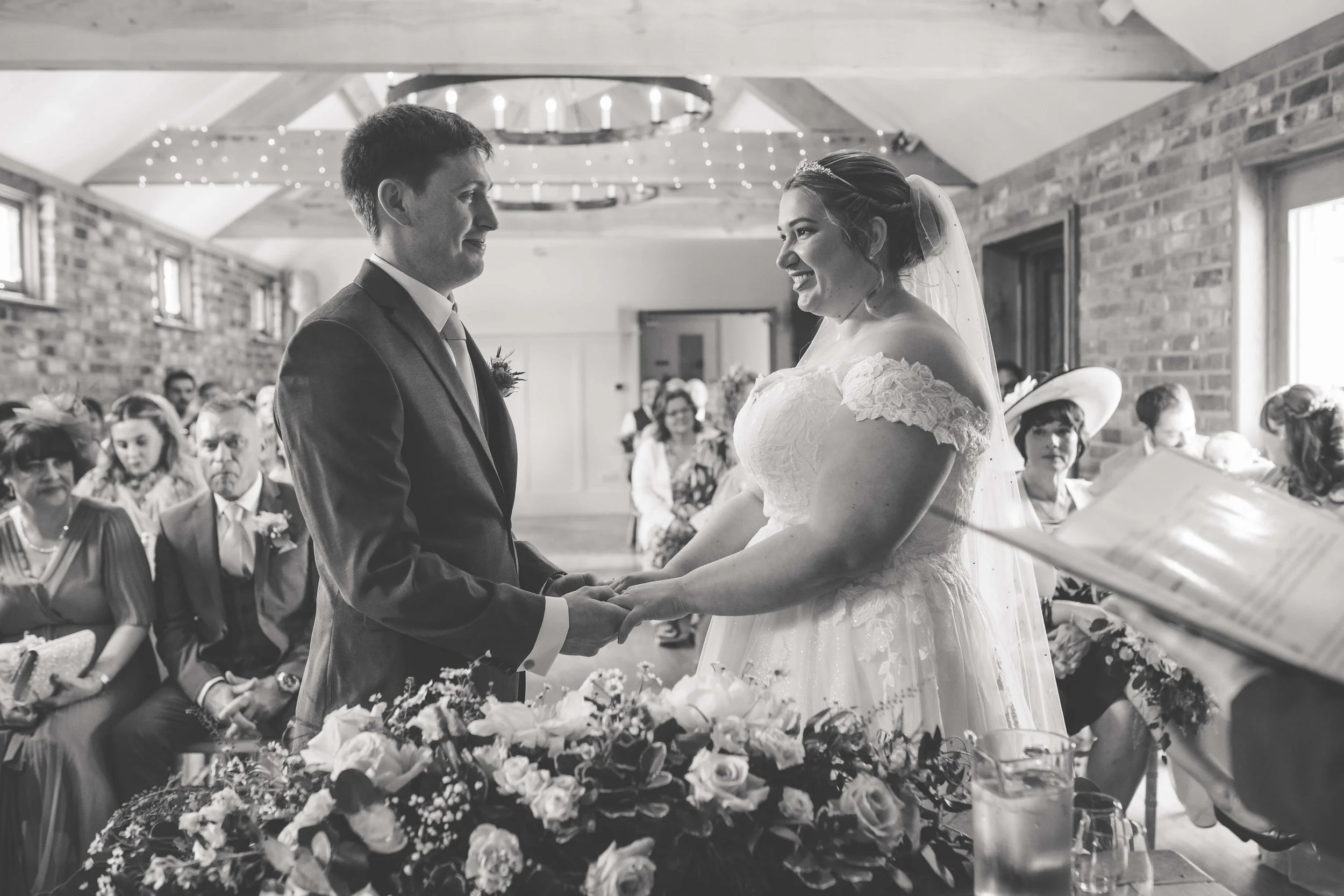 A bride and groom holding hands during a wedding ceremony in a rustic indoor venue with brick walls, surrounded by seated guests.