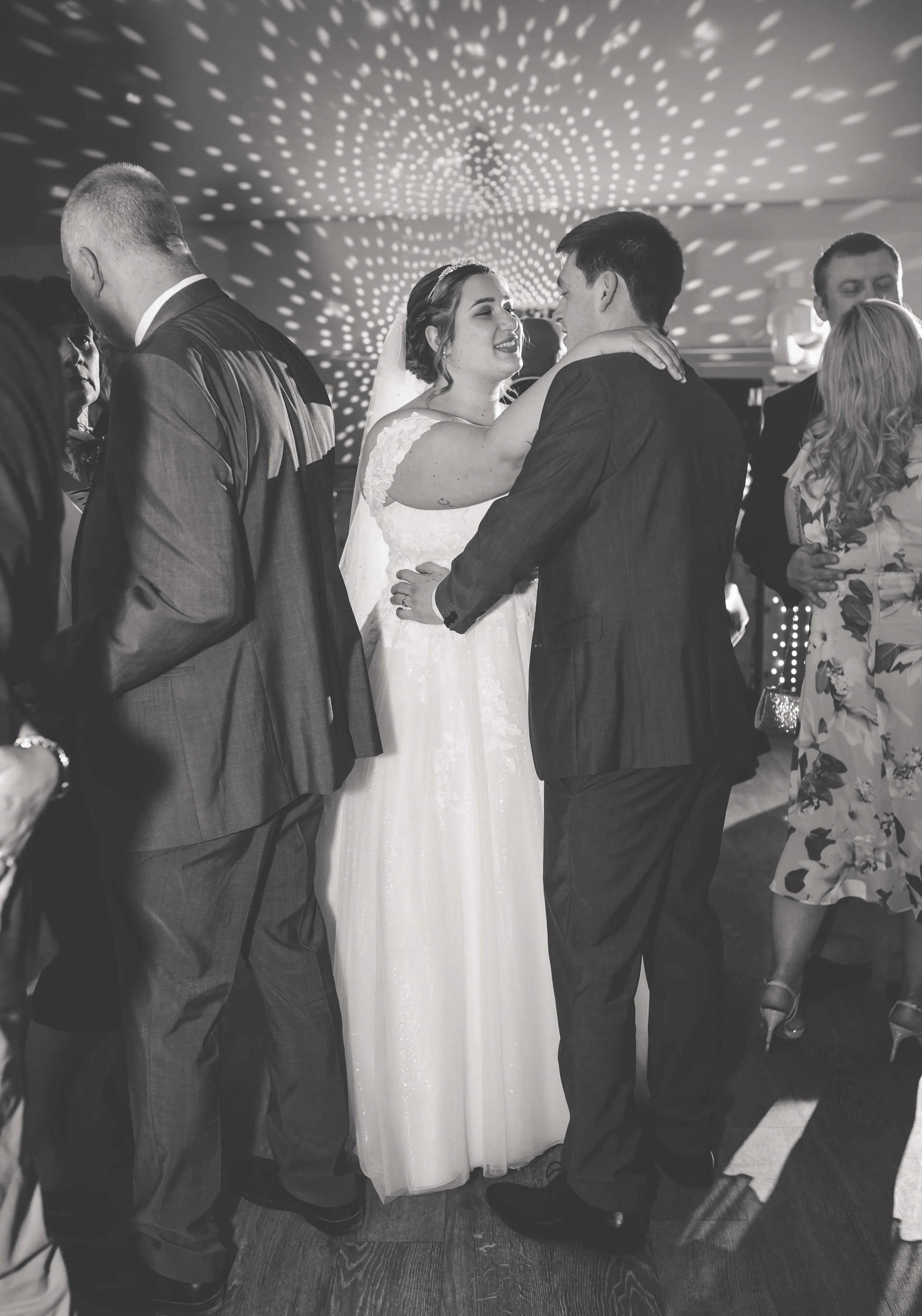 A bride and groom sharing a dance at their wedding reception, surrounded by guests. The setting has decorative lights on the ceiling.