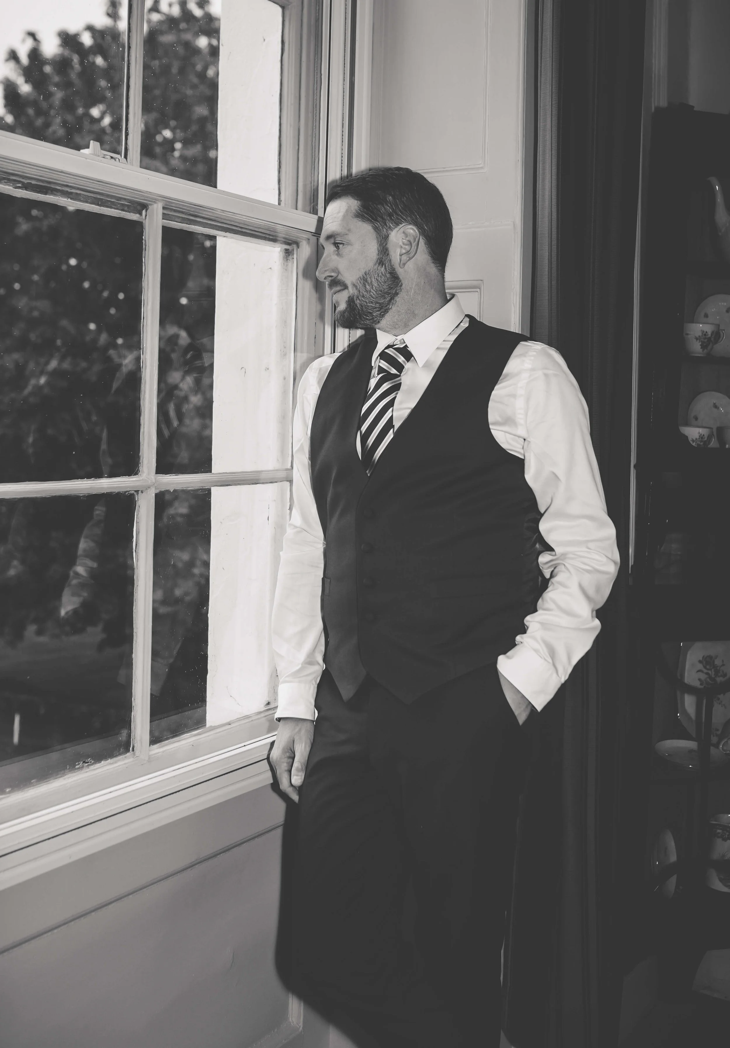 A man with a beard, dressed in a white shirt, striped tie, and vest, stands near a window looking outside with hands in pockets