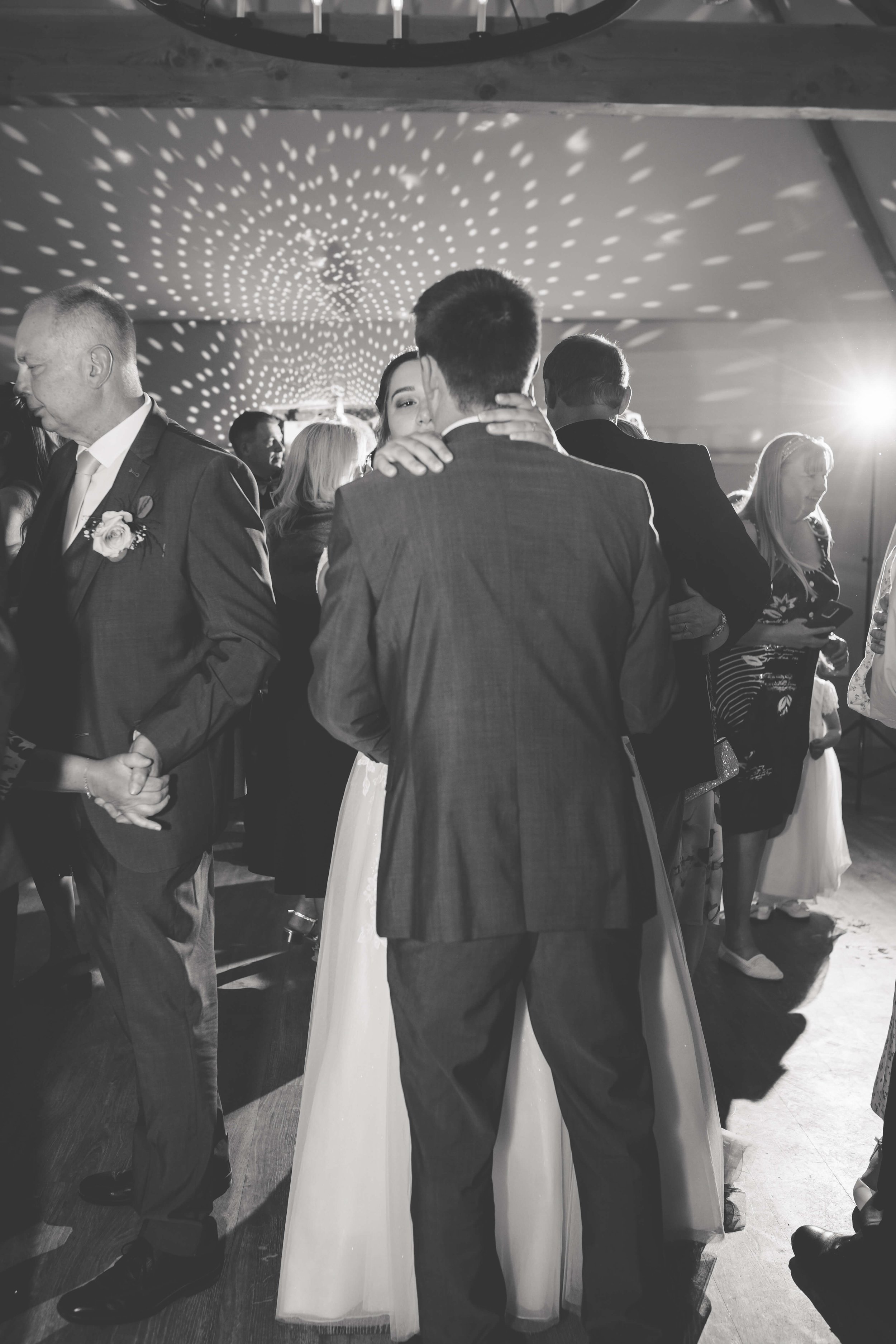A black and white photo of a wedding reception with couples dancing closely in the center, including a bride in a white gown and a groom in a tuxedo, under a ceiling with decorative lighting.