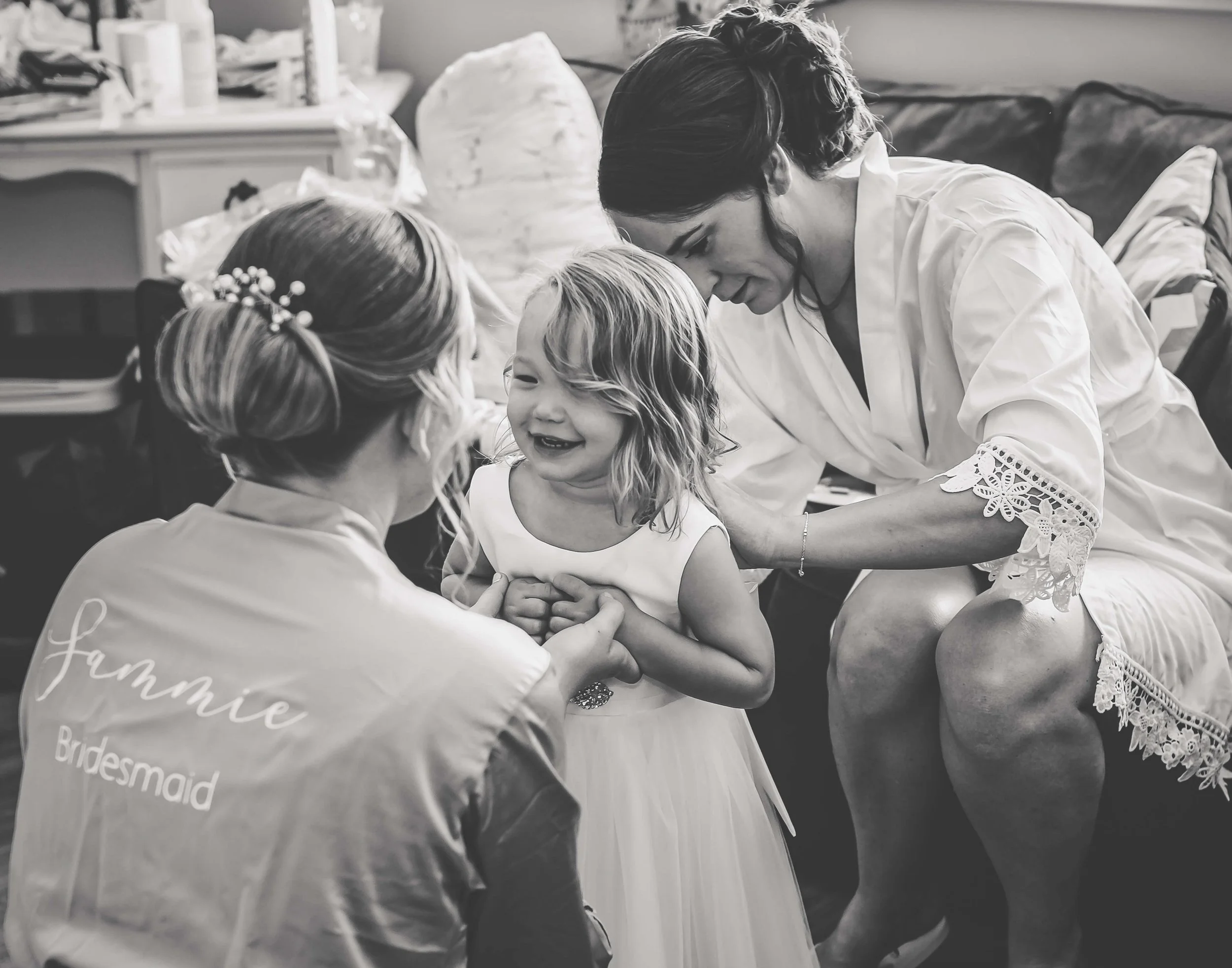A young girl dressed in a white dress is smiling and holding hands with a woman, likely her mother, while another woman dressed as a bridesmaid with 