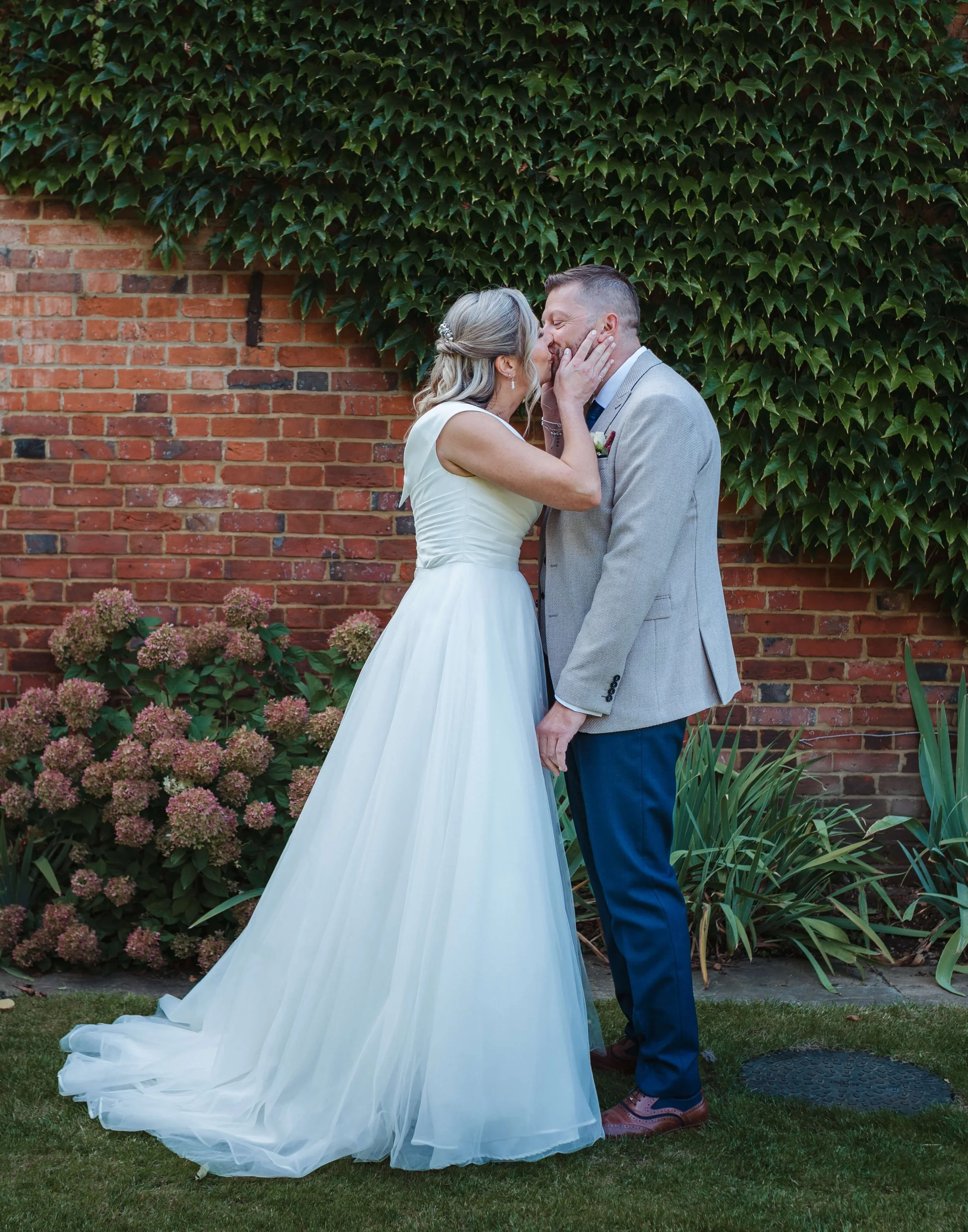 A bride and groom share a kiss outdoors in front of a brick wall covered with green ivy. The bride wears a white dress with a long train, and the groom wears a light gray suit jacket and blue pants.