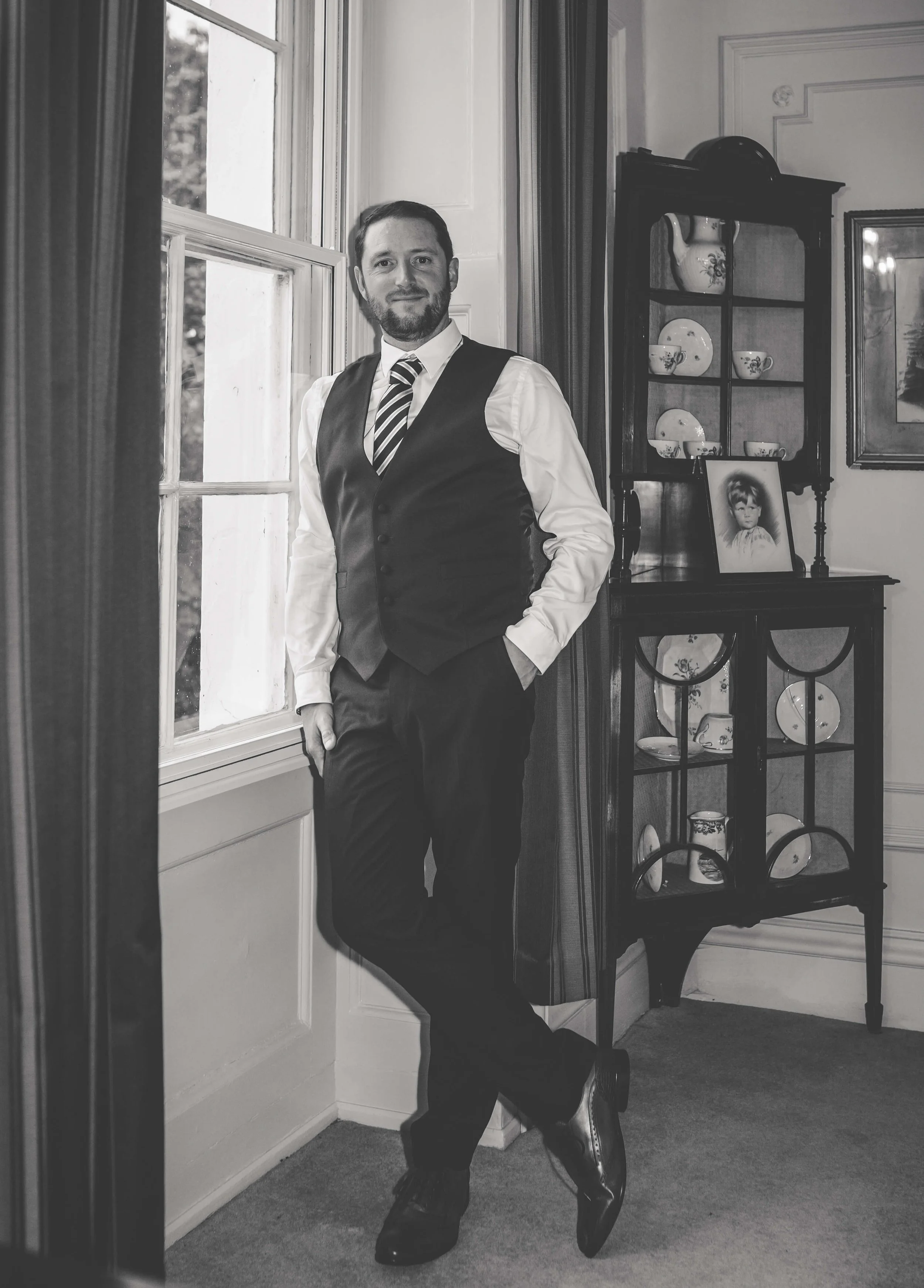 A man in formal attire, wearing a waistcoat, tie, and dress shoes, stands near a window in a room with vintage decor, including a china cabinet and framed pictures.