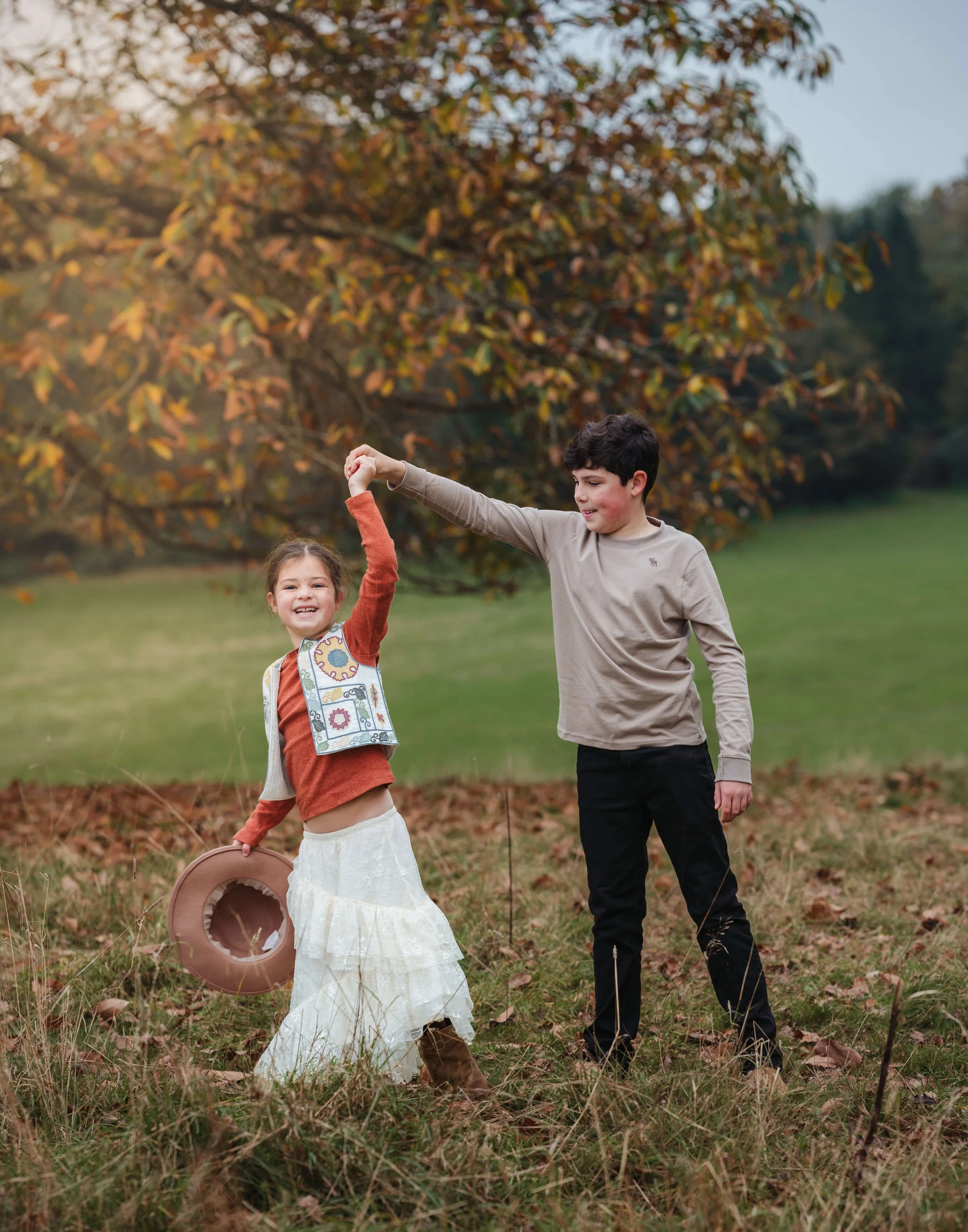 A boy and girl dancing outdoors in a park during autumn, holding hands. The girl has a floral vest, a white skirt, and brown boots, while the boy wears a beige long-sleeve shirt and black pants. There are autumn leaves on the ground and a tree with c