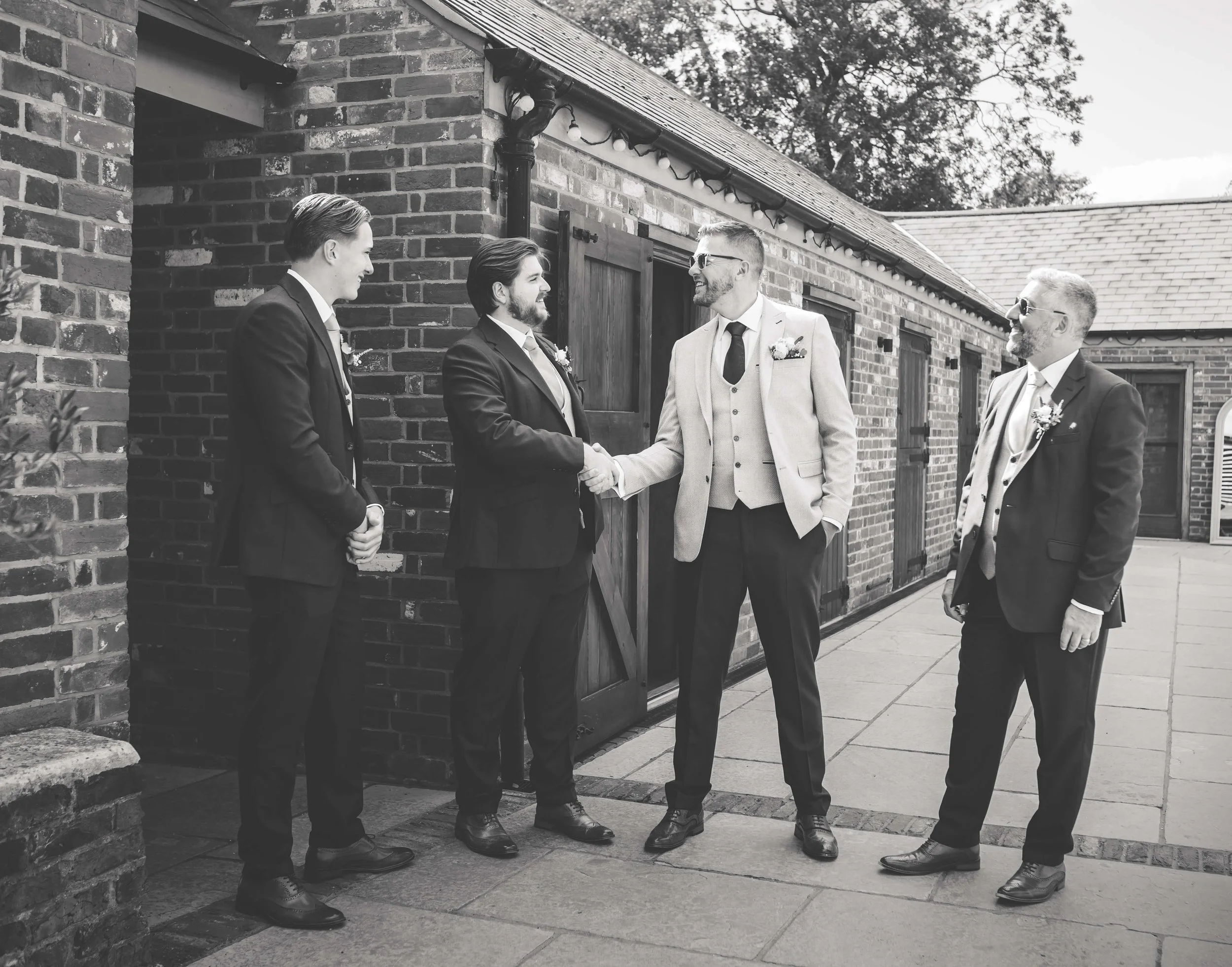 Five men in suits at a wedding, with one man shaking hands with another, on a paved outdoor area in front of a brick building.