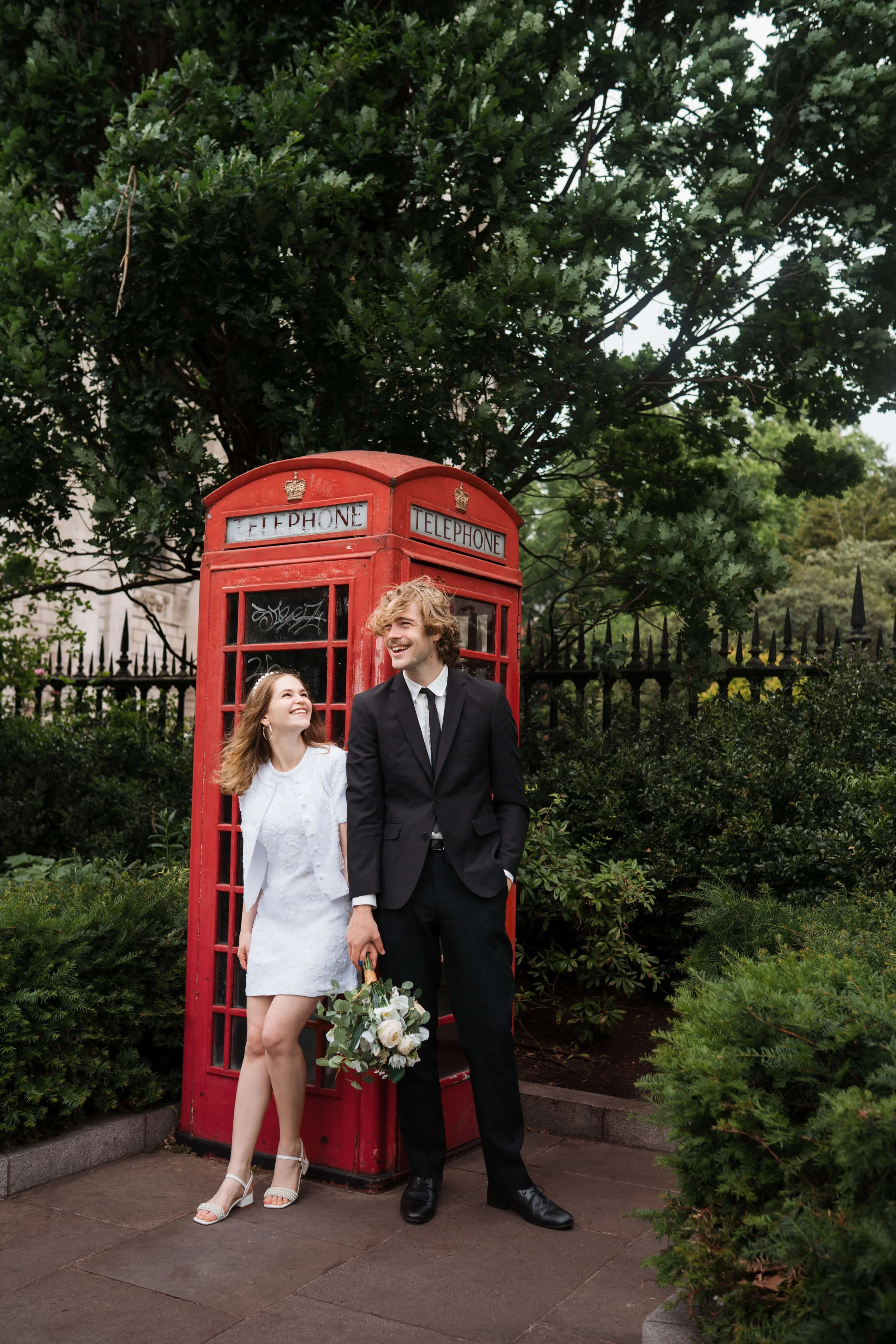A couple in wedding attire standing in front of a red British telephone booth, surrounded by greenery and smiling at each other.